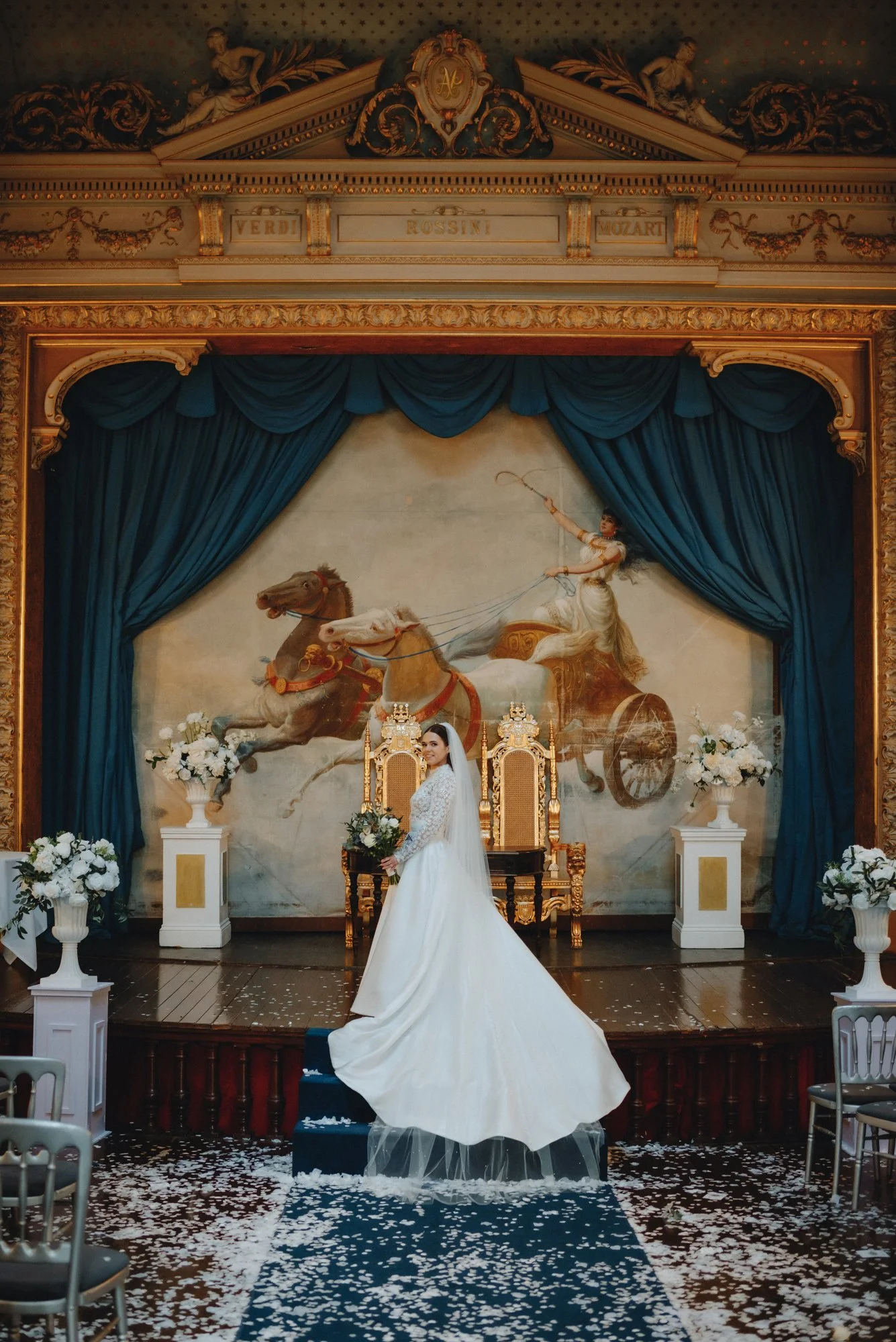 Bride standing on a small set of stairs, holding a bouquet, in front of a royal-like throne with a baroque backdrop featuring a mural of a woman on a chariot pulled by horses. The stage is decorated with white flowers in tall vases, and dark blue cur