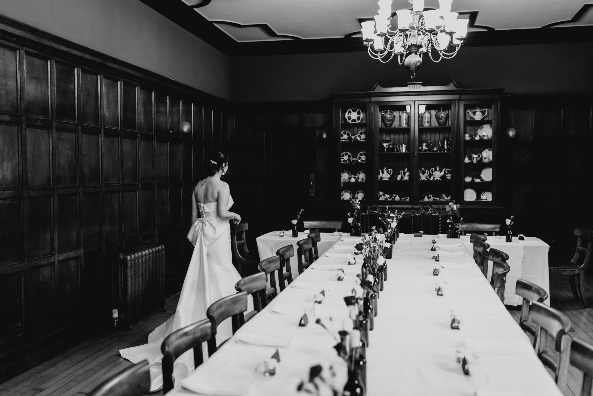 A woman in a wedding dress standing in a dimly lit dining room with a long table set for a gathering and a large china cabinet in the background.