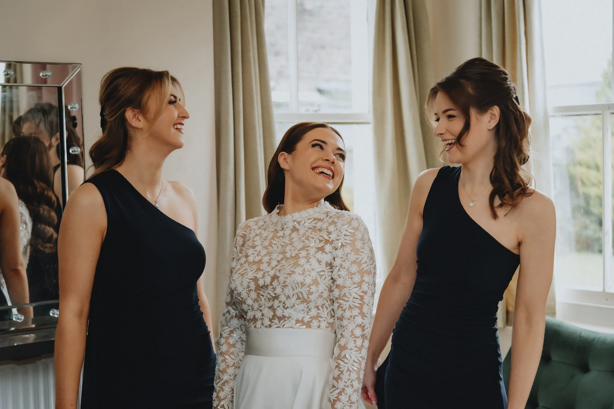 Three women smiling and engaged in conversation in a bright room with large windows and cream-colored curtains, one in a white dress with lace top, and two in black dresses.