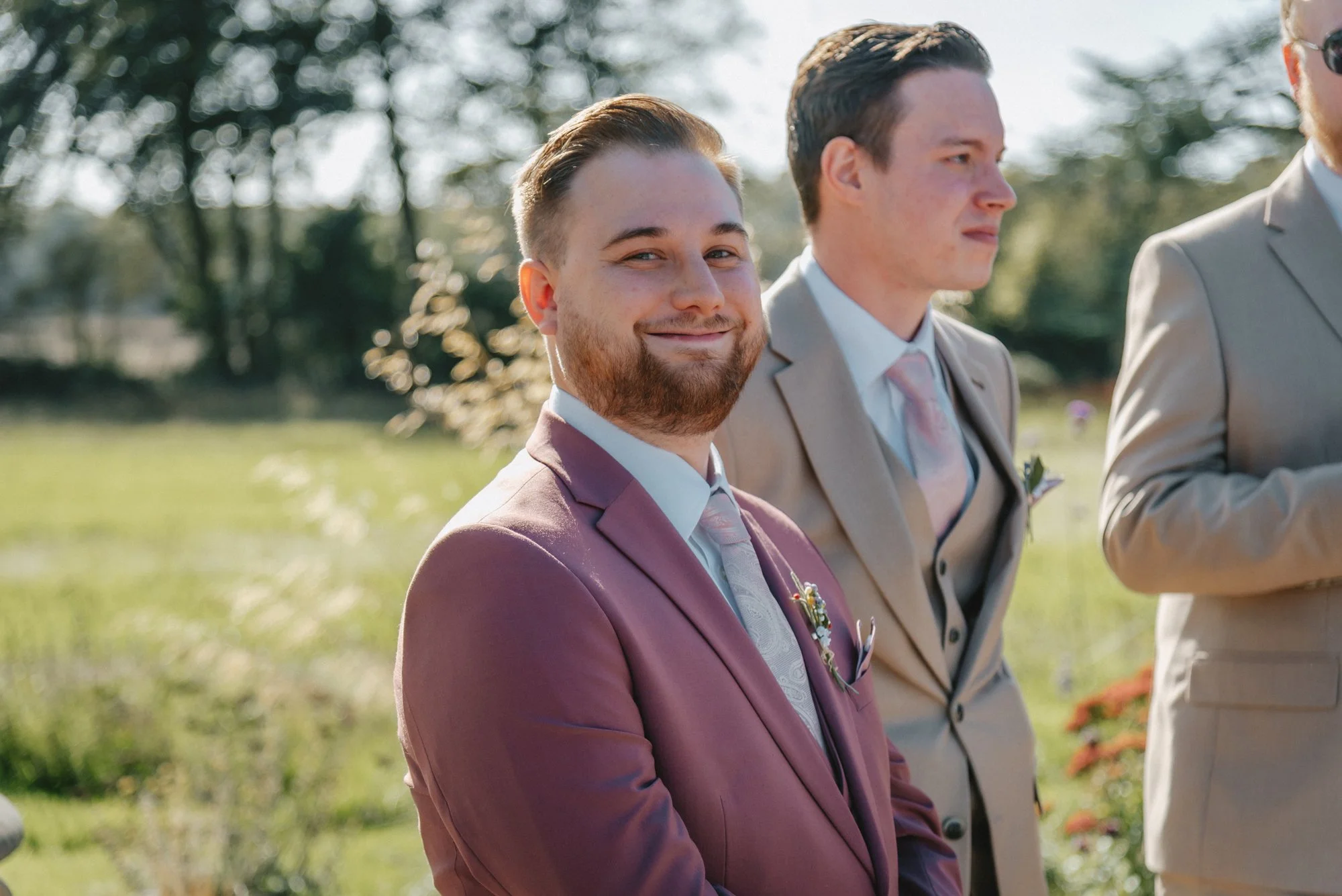 A man in a mauve suit smiling at a wedding outdoor ceremony, with another man in a beige suit next to him.