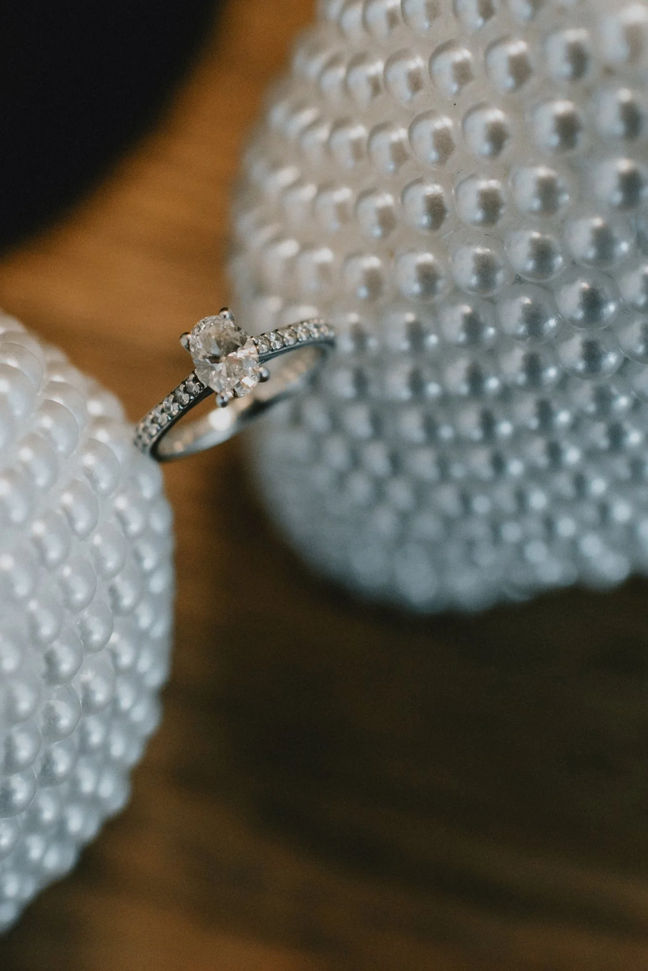 Close-up of an engagement ring with a heart-shaped diamond, surrounded by smaller diamonds on a band, placed between two textured pearl objects.
