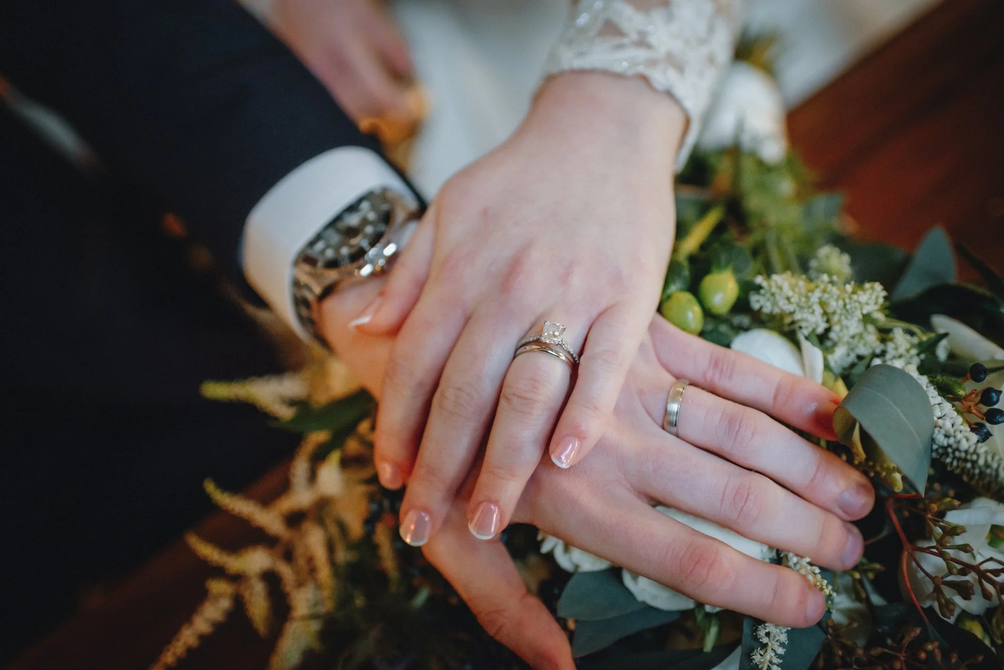 Close-up of a newlywed couple's hands with wedding rings, resting on a bouquet of white and green flowers.