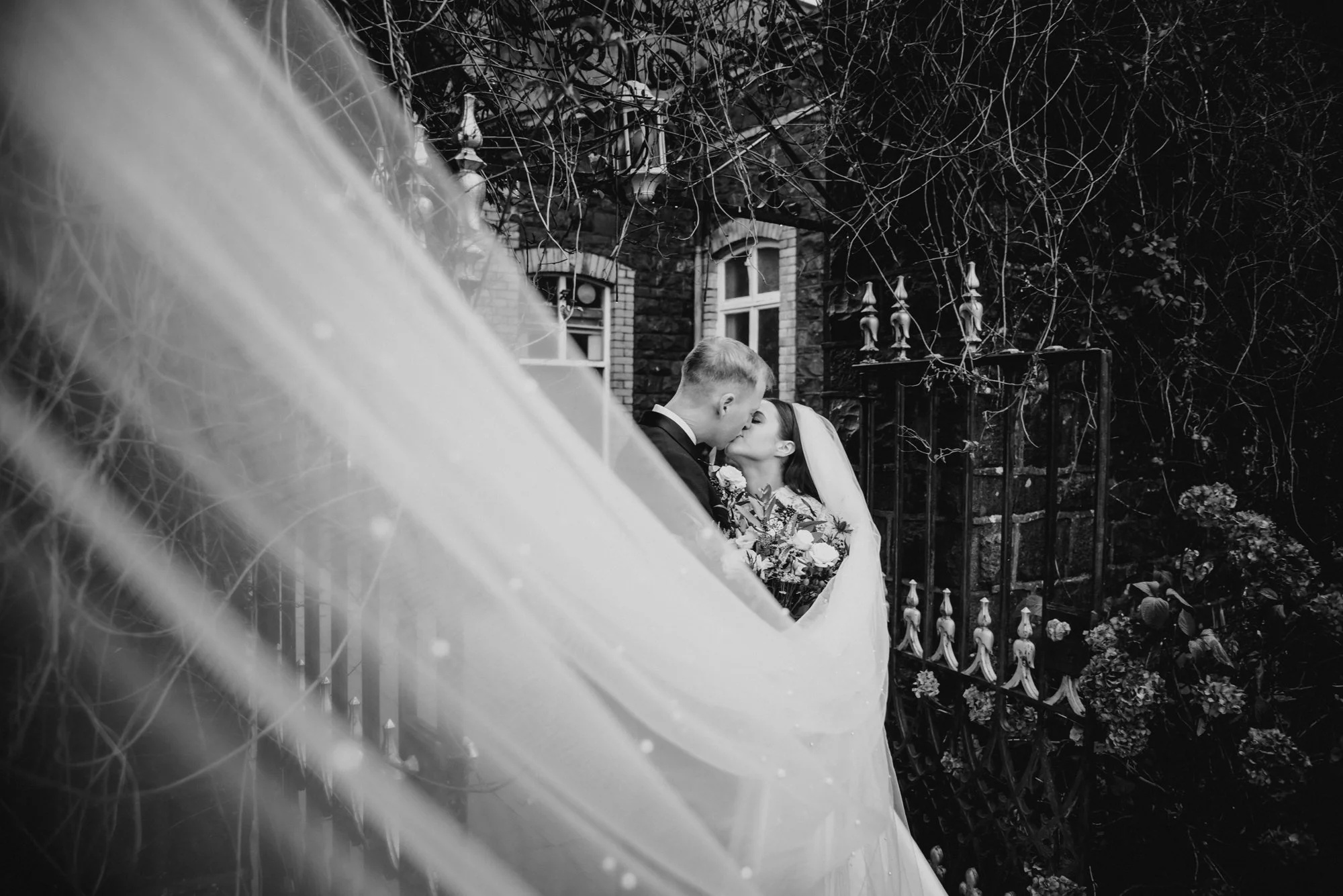 A black and white photo of a bride and groom sharing a kiss behind a veil. The bride holds a bouquet, and they are outside near a brick building with windows and decorative ironwork, surrounded by foliage.