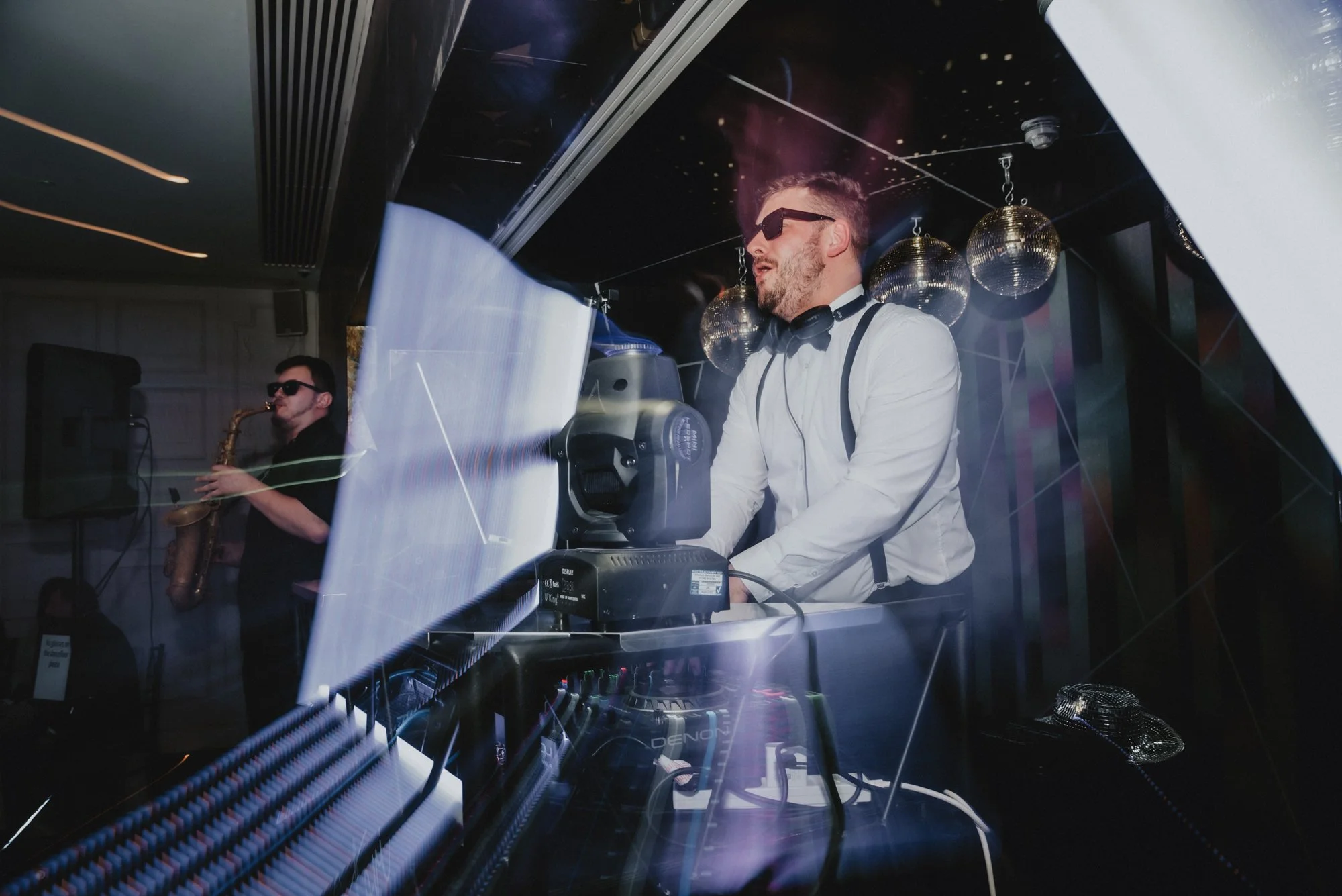 DJ with sunglasses at a nightclub with disco balls, music equipment, and a saxophonist in the background.