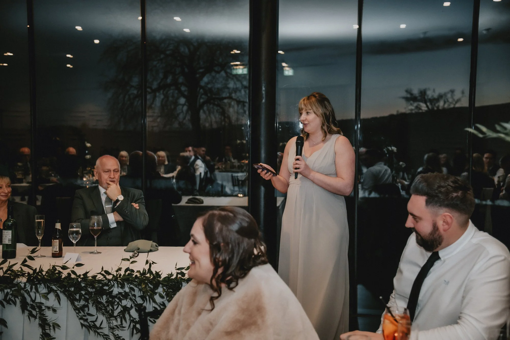 A woman in a beige dress giving a speech with a microphone at a formal event, seated guests looking on, with a window showing a dark outdoor scene in the background.