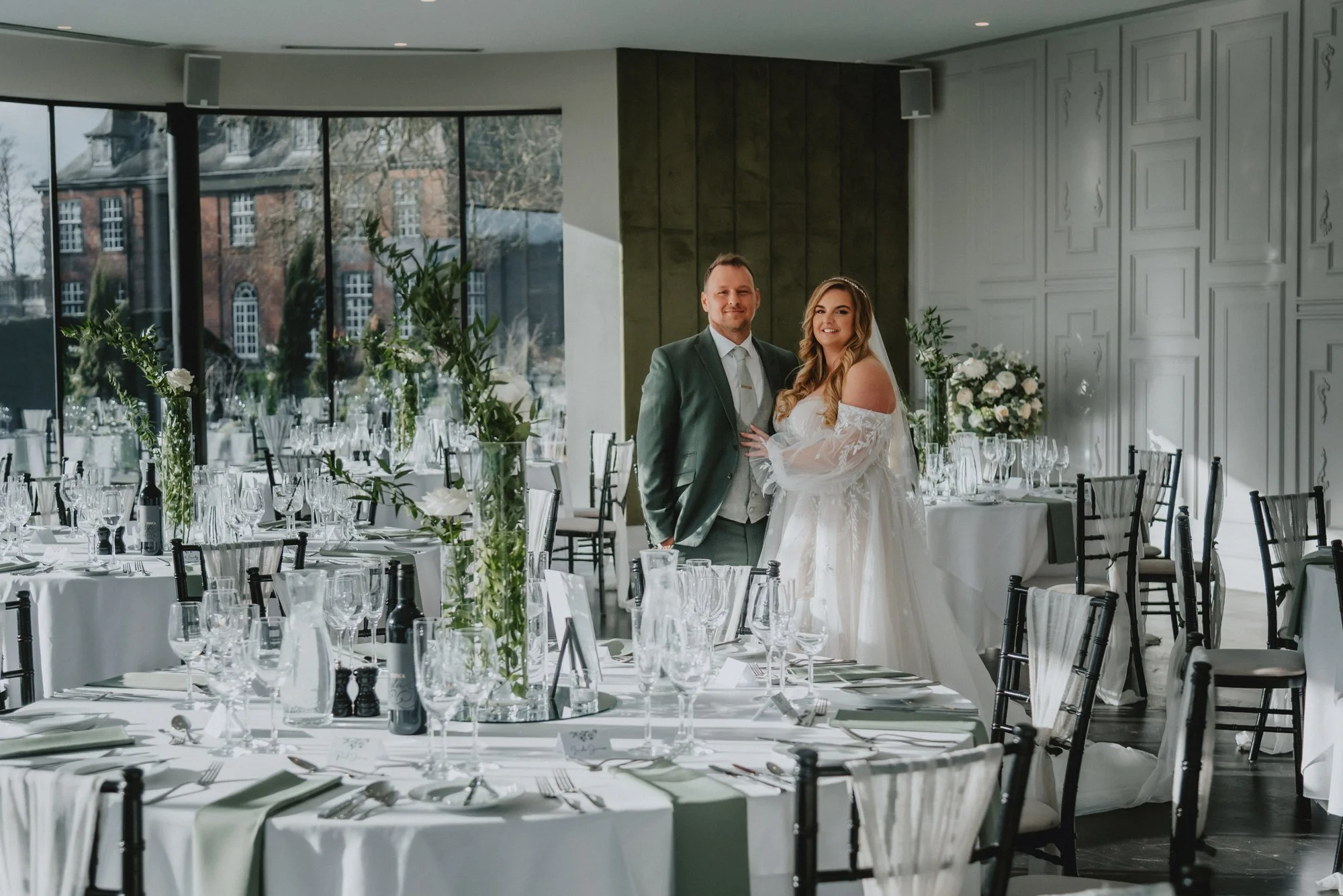 A newlywed couple standing in a beautifully decorated wedding reception hall with round tables set with glassware, cutlery, and white tablecloths, featuring tall floral centerpieces and large windows in the background.