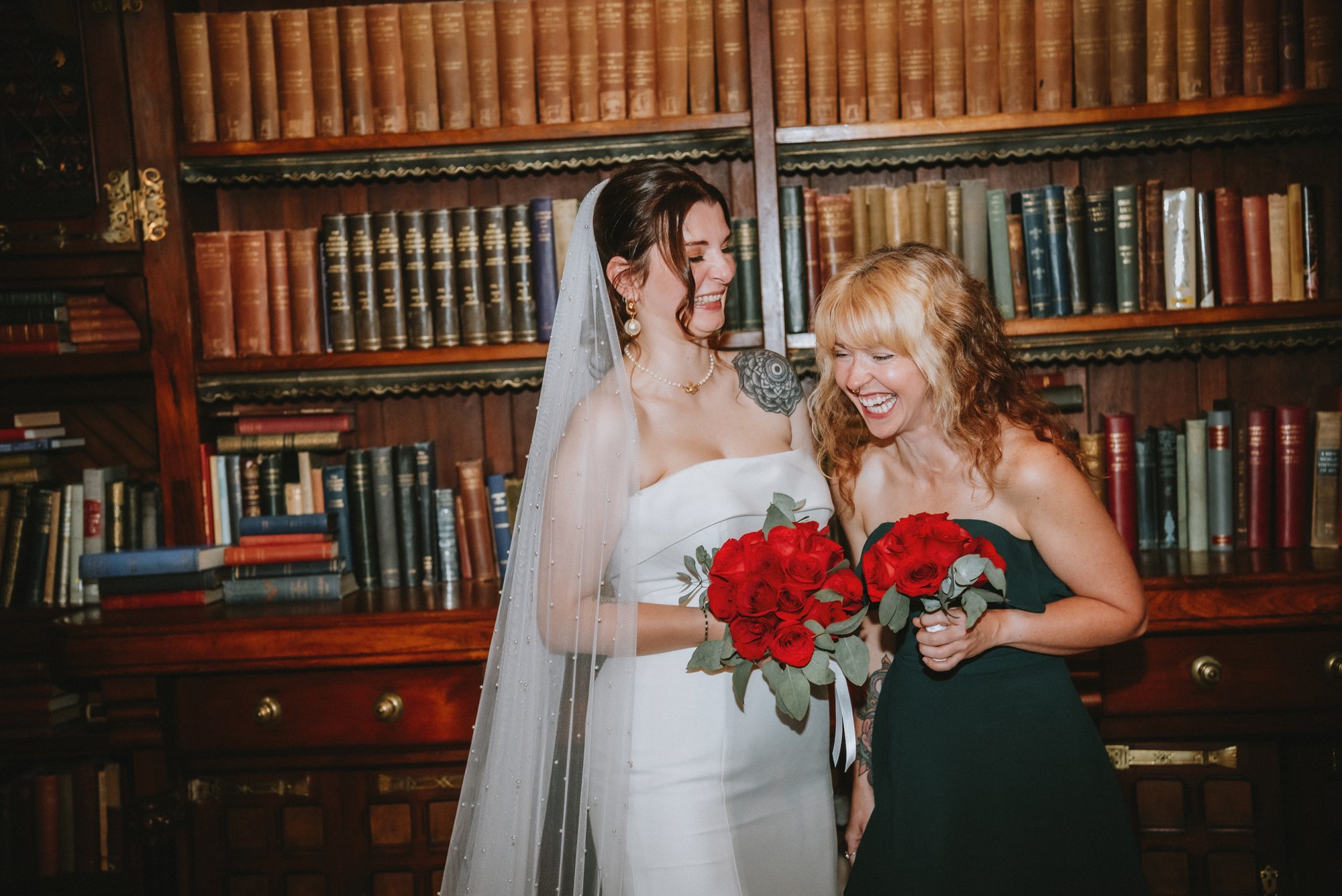 Two women smiling and holding bouquets of red roses, standing in front of a wooden bookshelf filled with books.