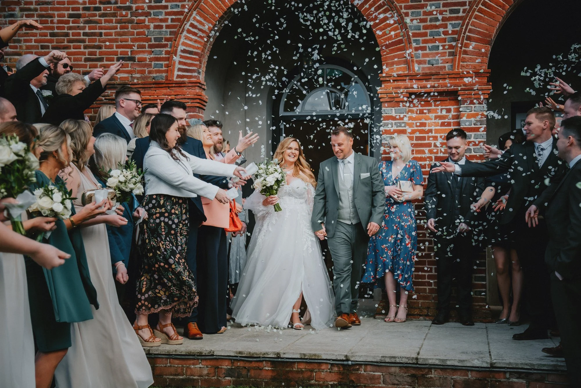 A bride and groom holding hands and walking outside a brick building, surrounded by celebrating guests throwing confetti at their wedding