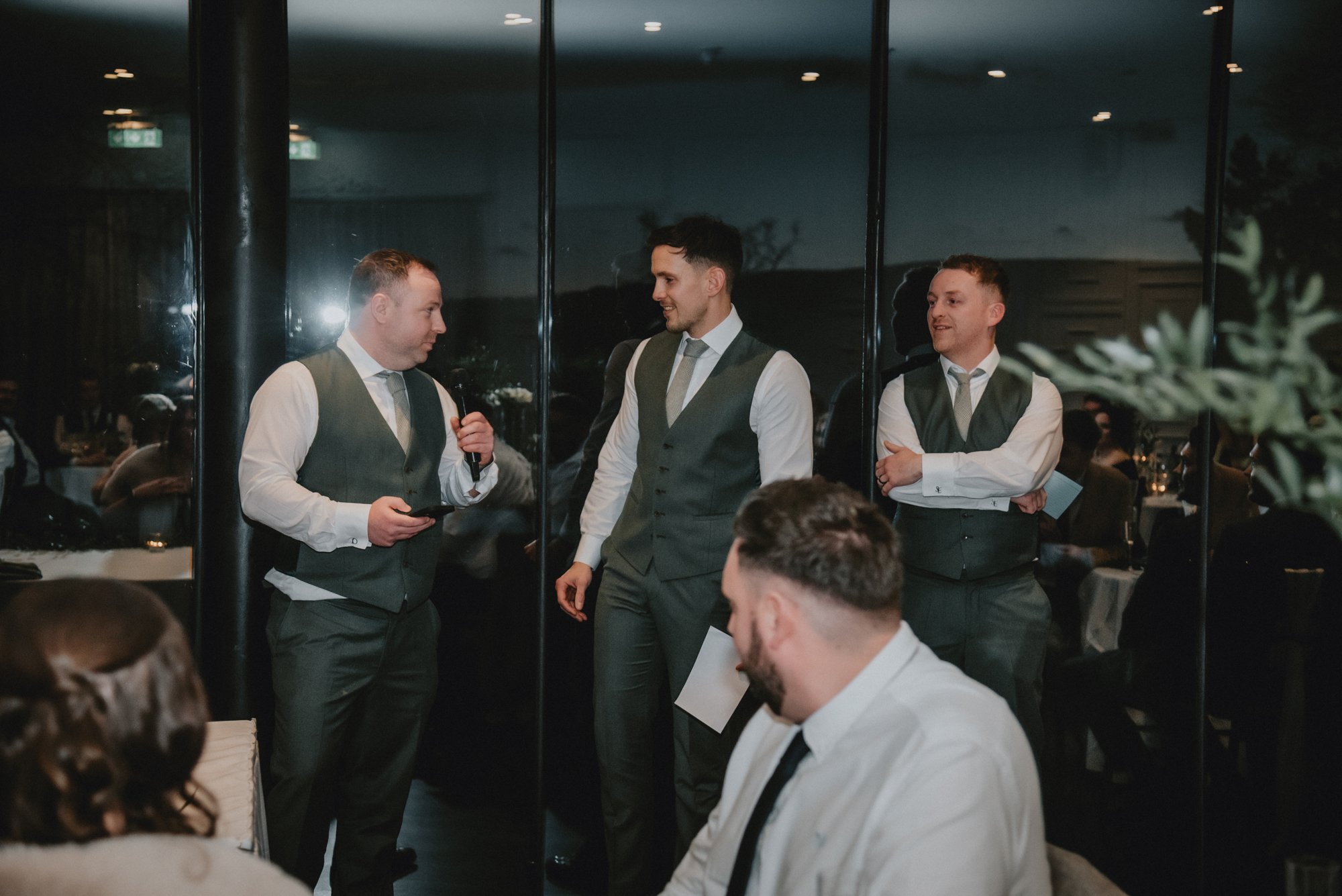Three men in formal suits and vests standing and talking at an indoor event, with a dark background and reflections on the large glass window behind them. Another person is seated in the foreground.
