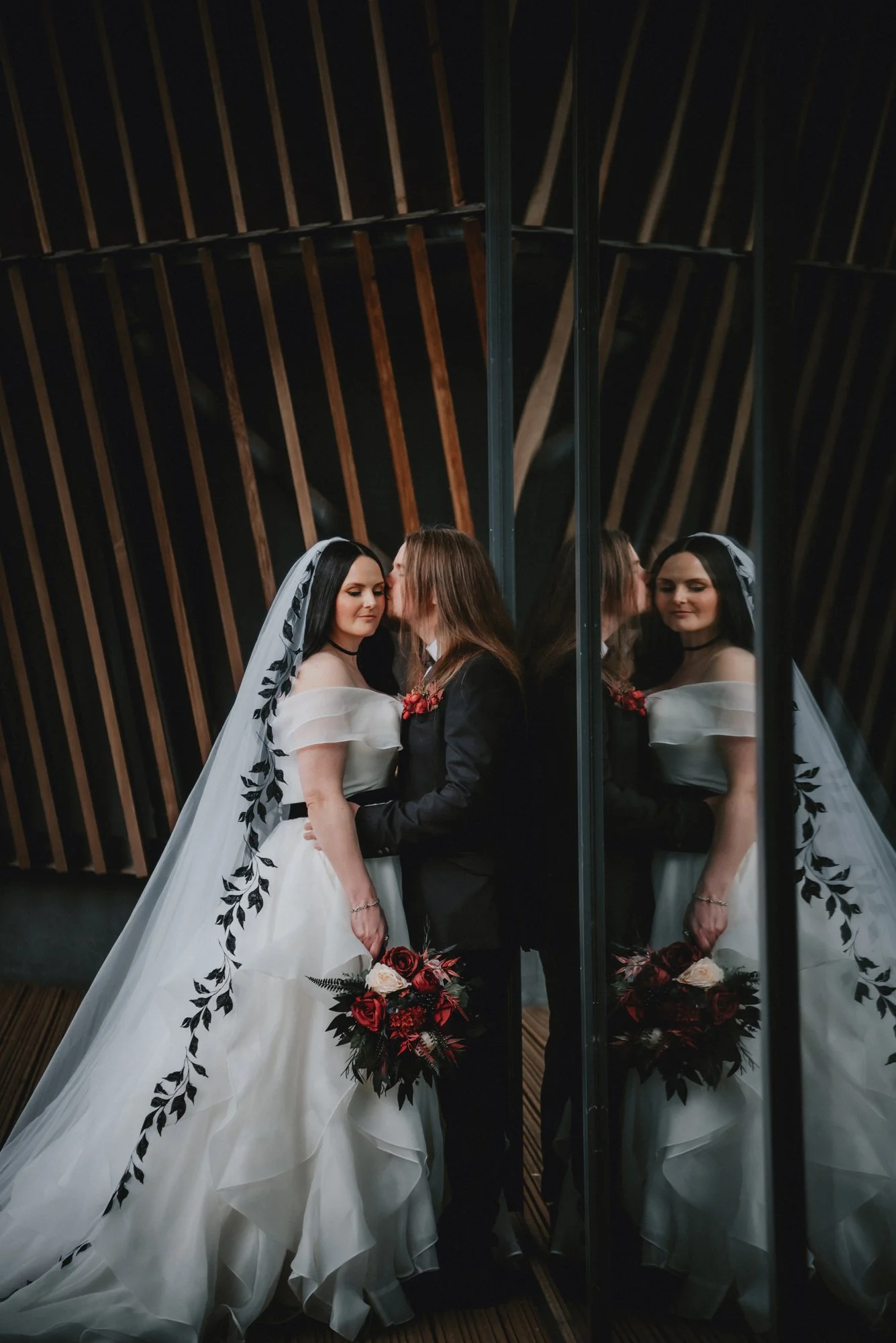 Wedding couple, bride in a white dress and veil, and groom in a black suit, standing close with eyes closed, holding a bouquet of red and white flowers, reflected in a mirror.