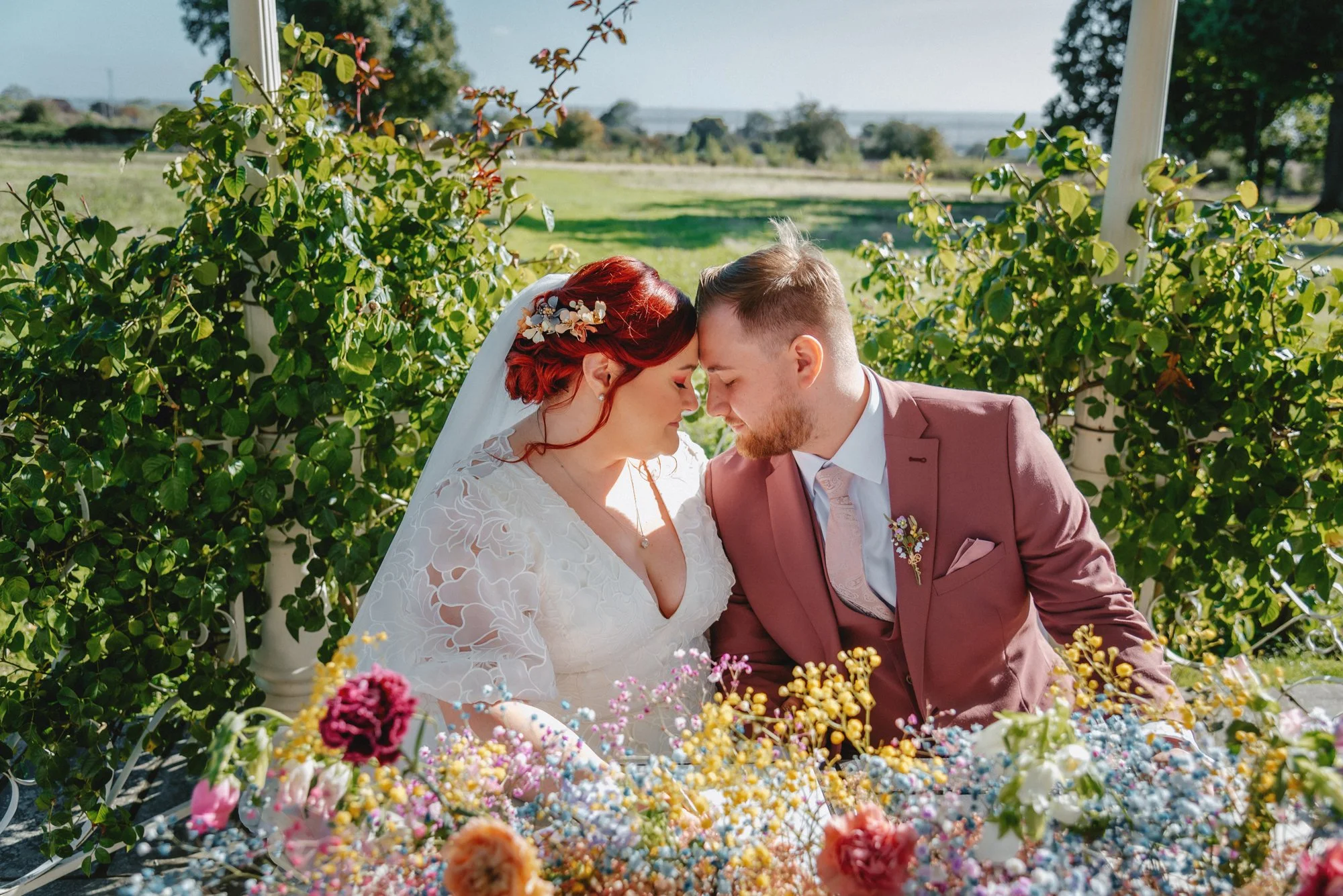 A bride and groom sit closely together outdoors, forehead to forehead with eyes closed, surrounded by lush greenery and colorful flowers.