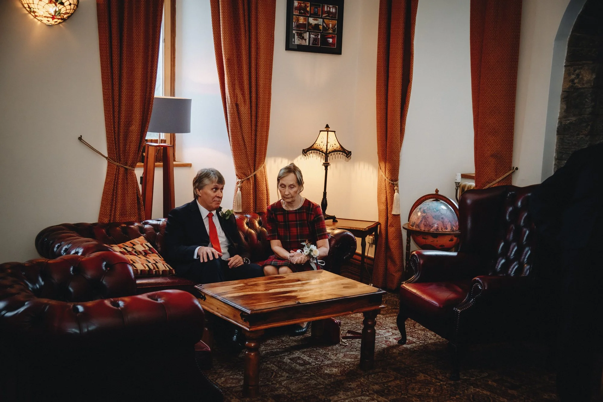 Two elderly women sitting on a dark red leather sofa in a cozy room with warm lighting, orange curtains, a wooden coffee table, a globe, a framed photo collage, and vintage lamps.