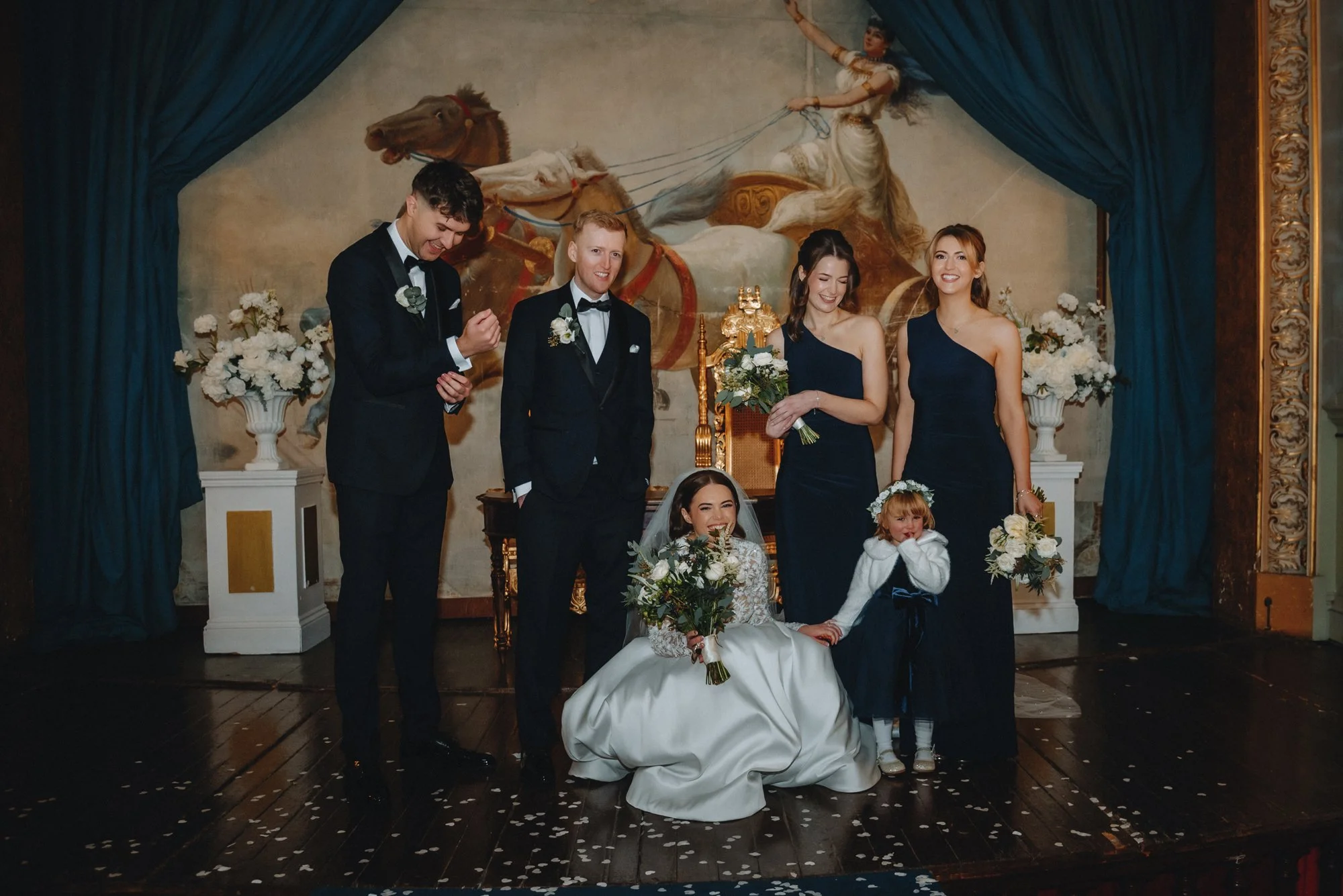 Group of wedding guests and a bride celebrate on a decorated stage with a mural backdrop, flowers, and curtains.