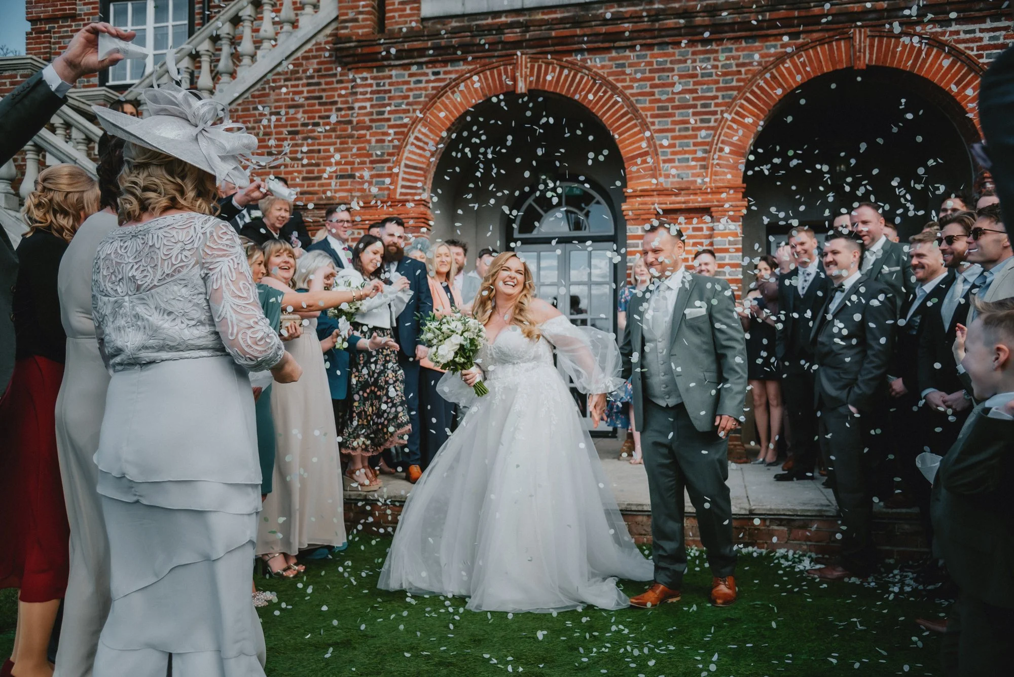 A joyful bride and groom celebrating their wedding with guests outside a brick building, confetti in the air.