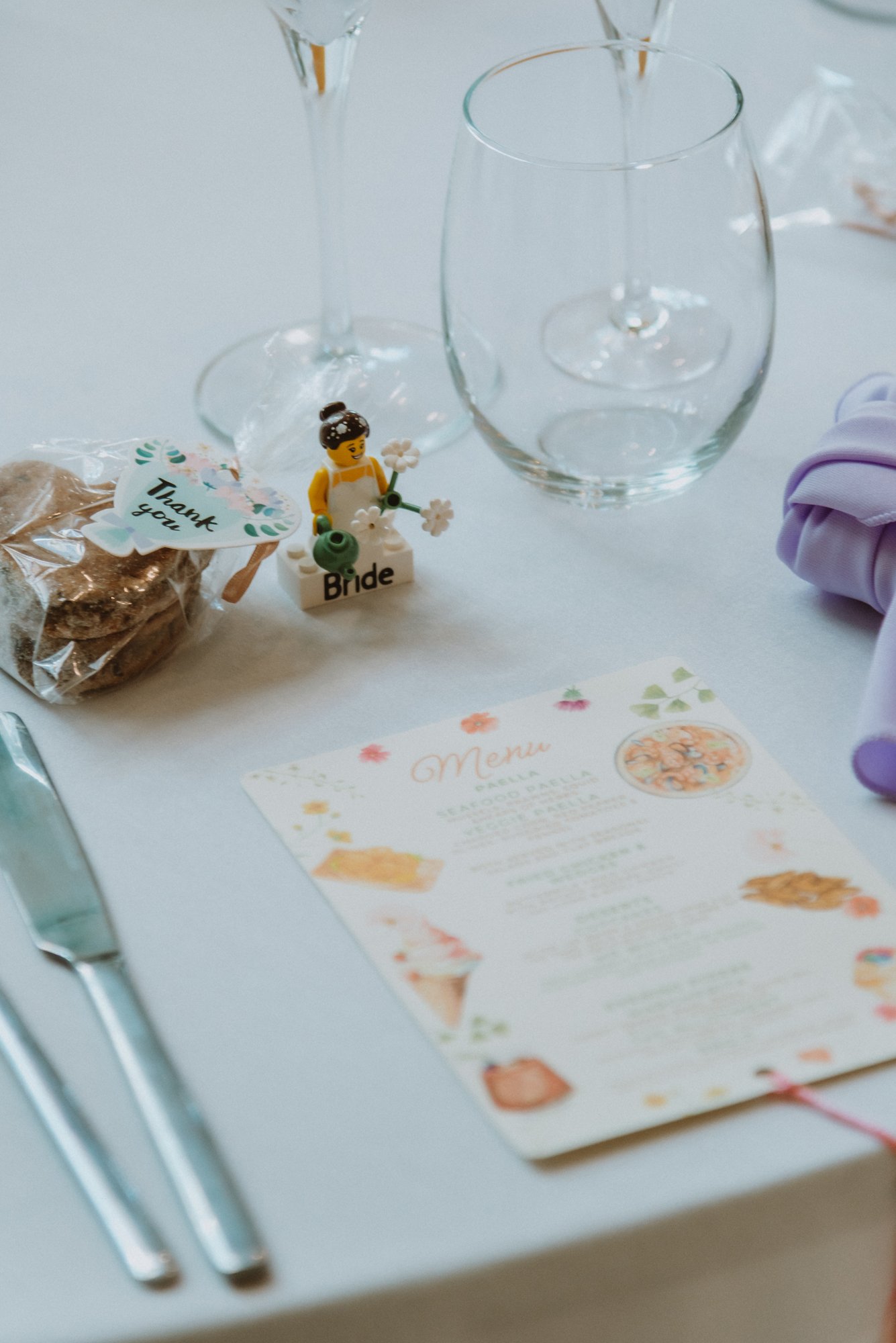 Table setting at a wedding with a toy bride figurine, a pink and purple napkin, a menu, and glasses for drinks.