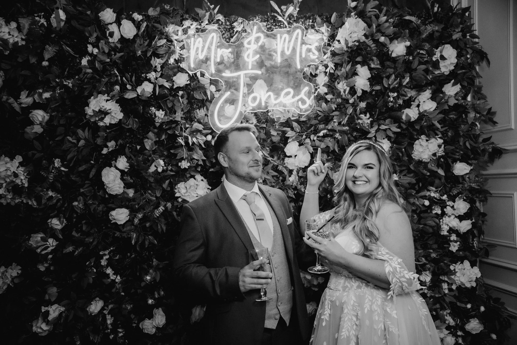 A bride and groom at their wedding reception, standing in front of a floral backdrop with a hanging sign that says 'Mr. & Mrs. Jones.' The bride is smiling and pointing upward, holding a drink, while the groom is looking at her and also holding a dri