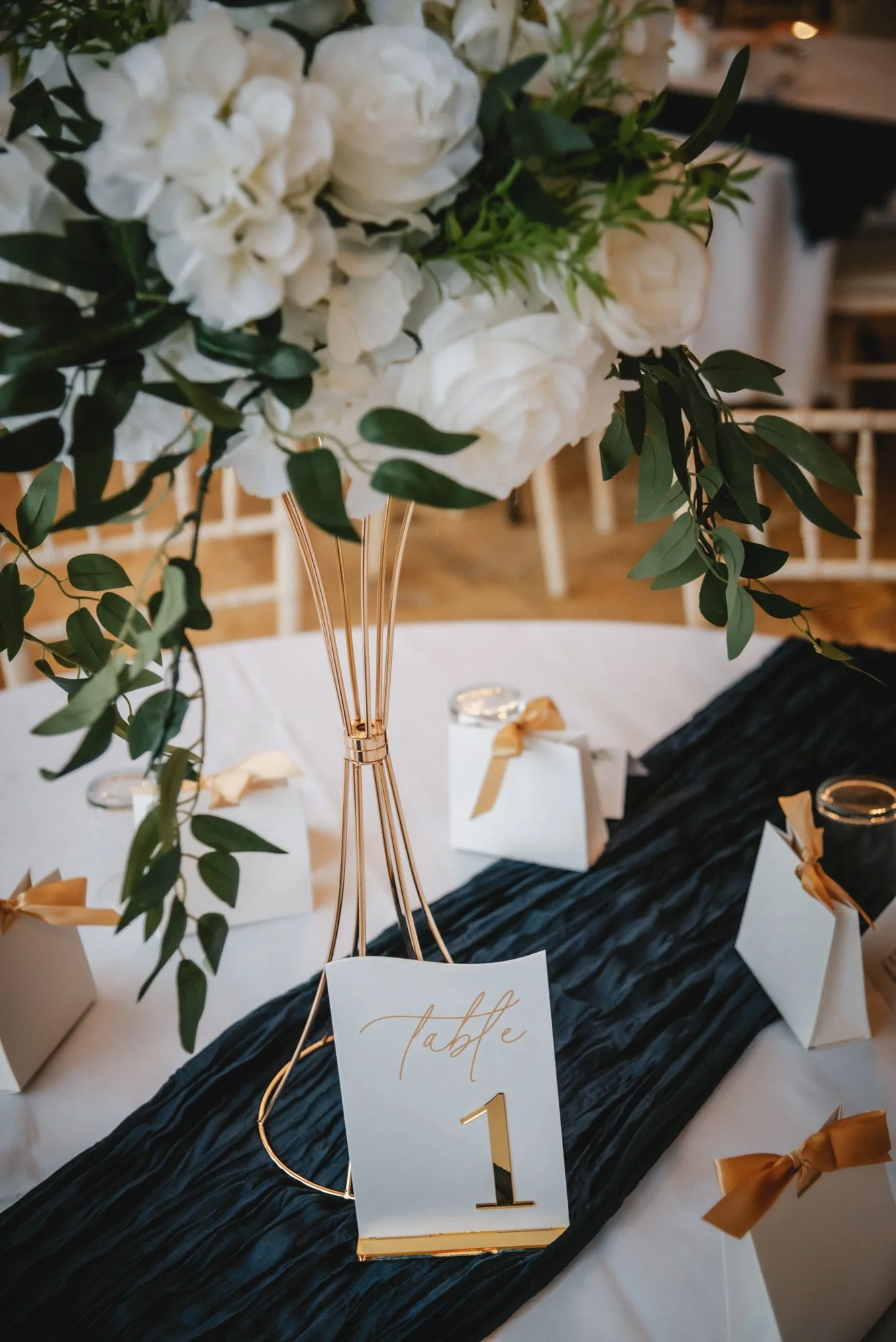 Table centerpiece with white flowers and green foliage in a tall gold vase, table number card indicating 'Table 1', surrounded by small white gift boxes with gold ribbons on a black table runner at a formal event.