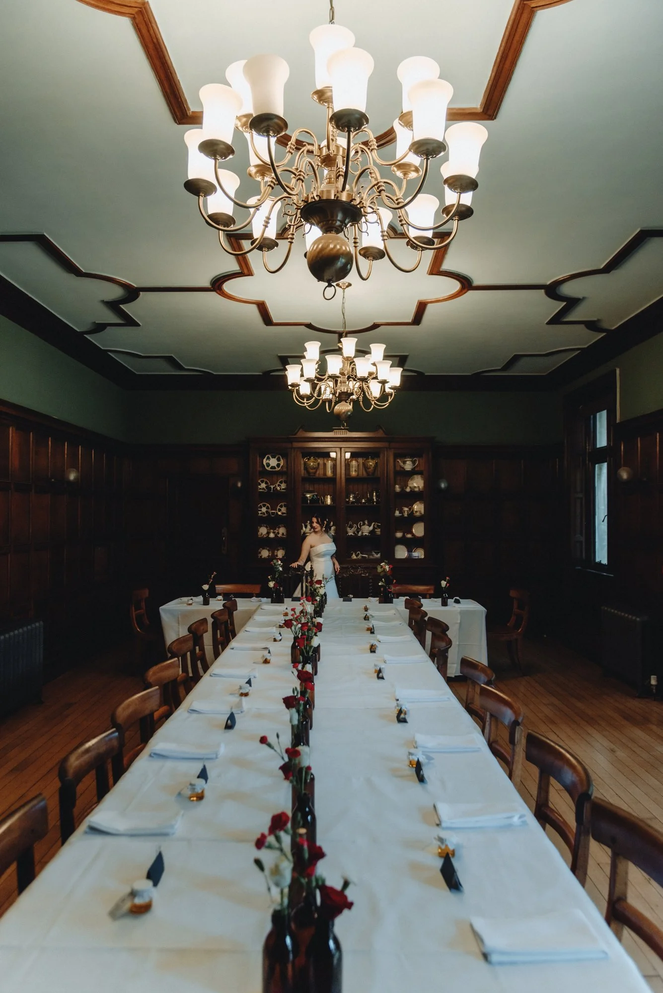 A formal dining room with a long table set for a meal, decorated with vases of red and white flowers, white napkins, and blue place cards. The room has dark wood paneling, two large chandeliers hanging from the ceiling, and a cabinet with china and g