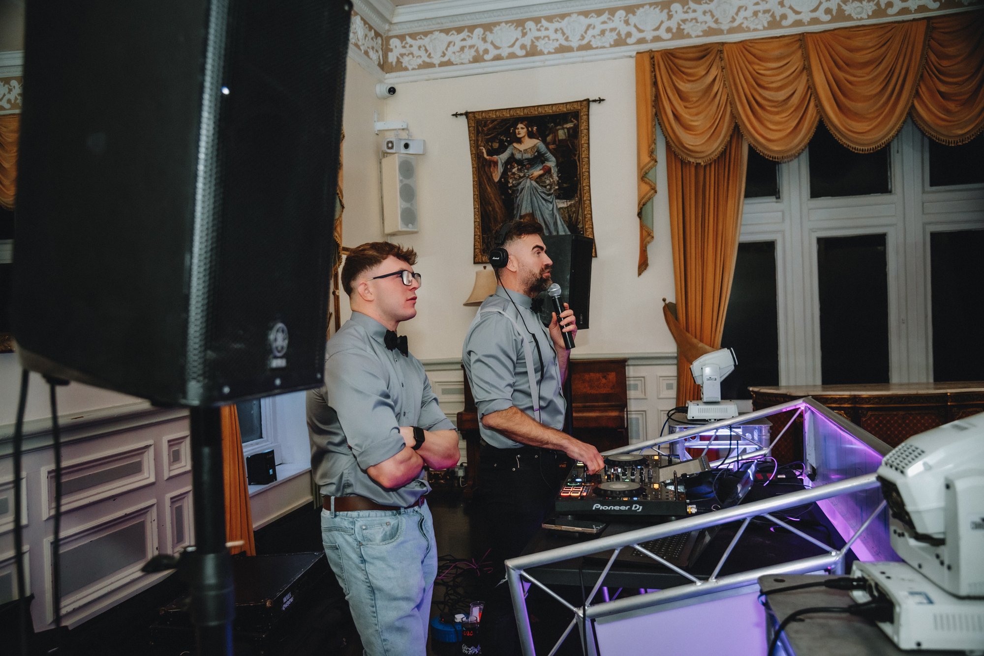 Two DJs, a man and a woman, are performing in a decorated room with gold curtains and ornate wall moldings. The man is speaking into a microphone with DJ equipment, while the woman stands beside him with arms crossed, both dressed in gray shirts and 