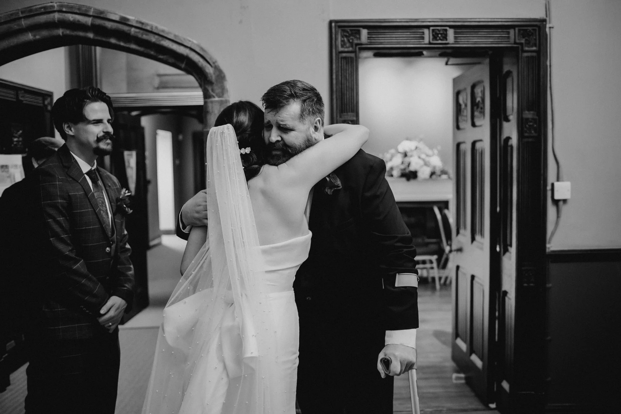 A bride hugging a man, likely her father, at a wedding in an indoor setting. Man has a cane and is dressed in a suit. A groom and another man are standing nearby, watching the scene. The image is in black and white.