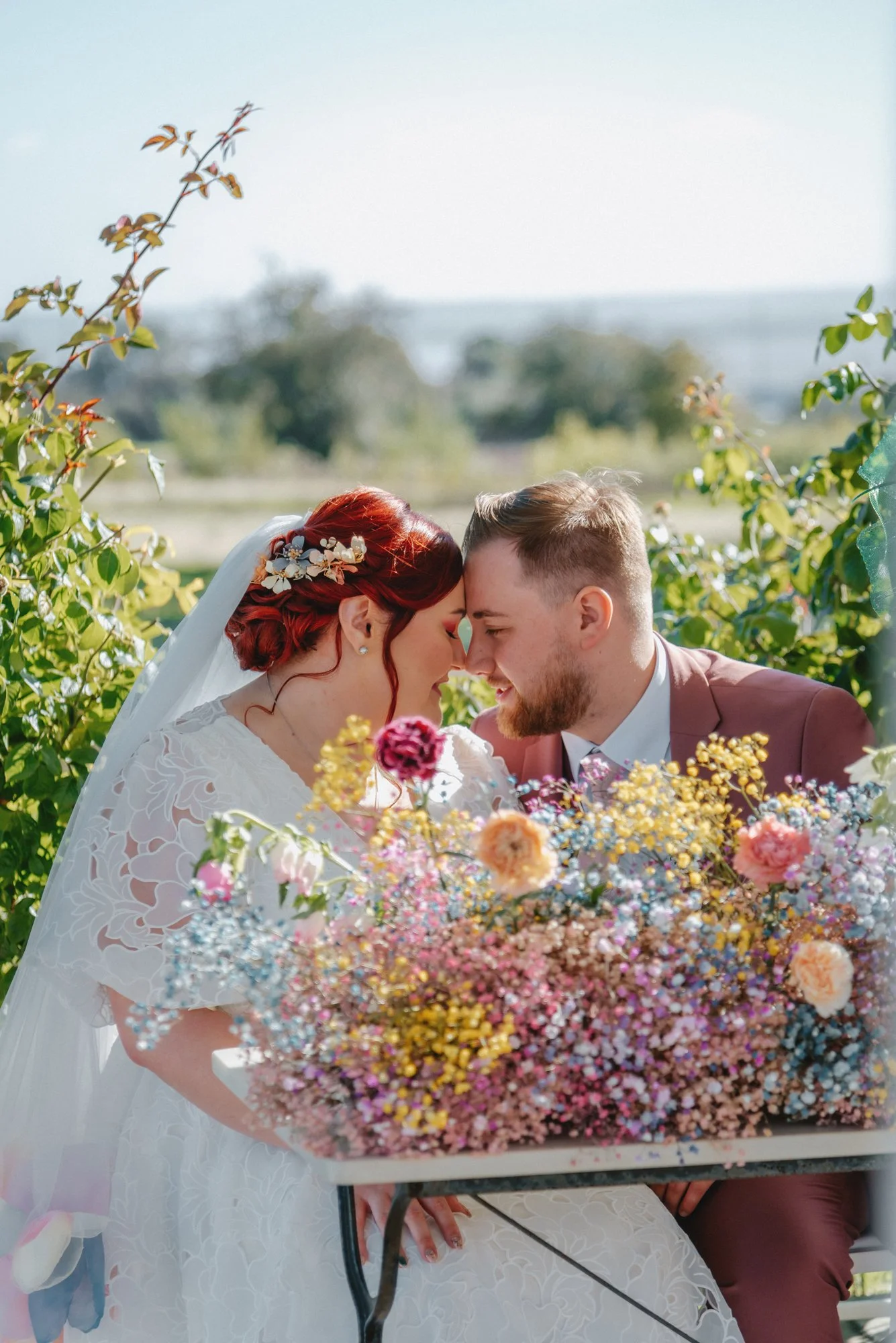 A bride and groom are touching foreheads and smiling, surrounded by colorful flowers during their wedding outdoor.