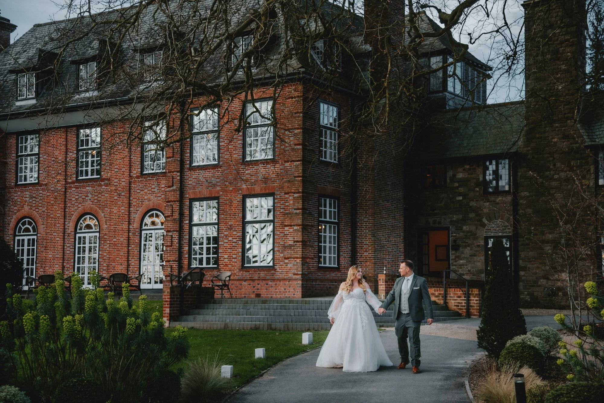 A bride and groom holding hands and walking outside a historic brick building during their wedding. The bride is in a white gown and the groom in a grey suit. The scene is set in a landscaped area with grass, plants, and trees.
