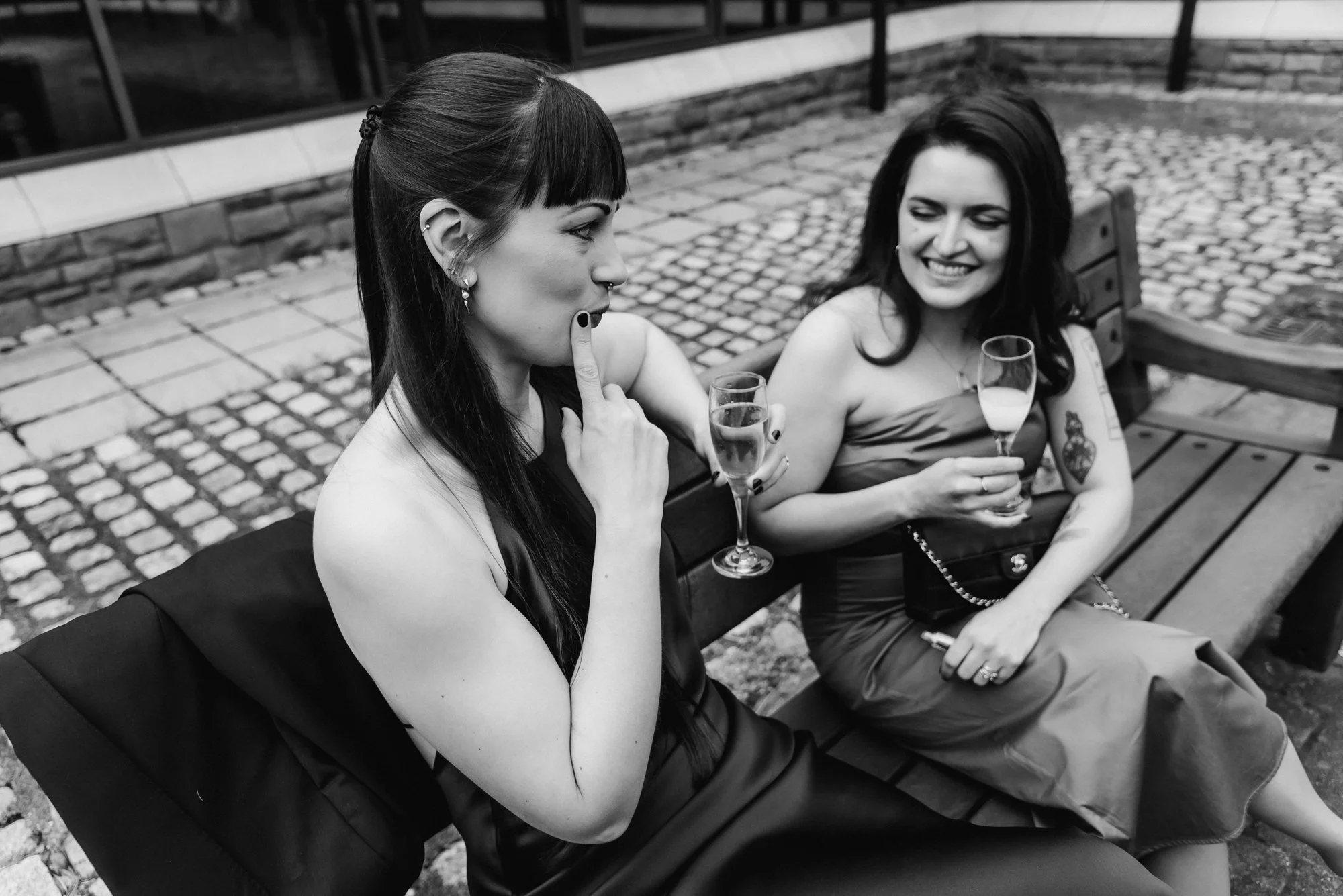 Two women sitting on a bench outside, smiling and talking while holding glasses of wine.