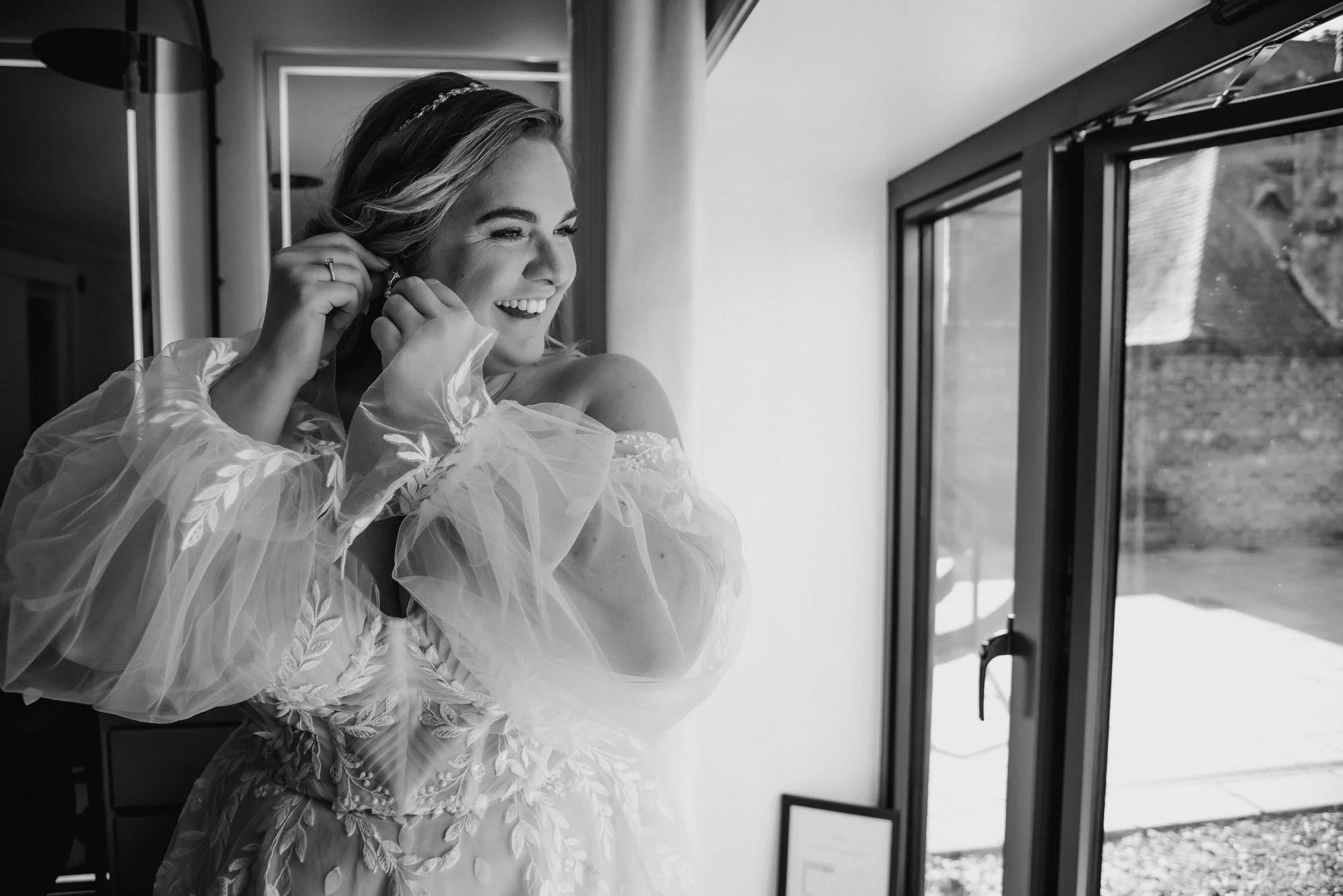 A woman in a wedding dress smiles as she puts on earrings by a window.