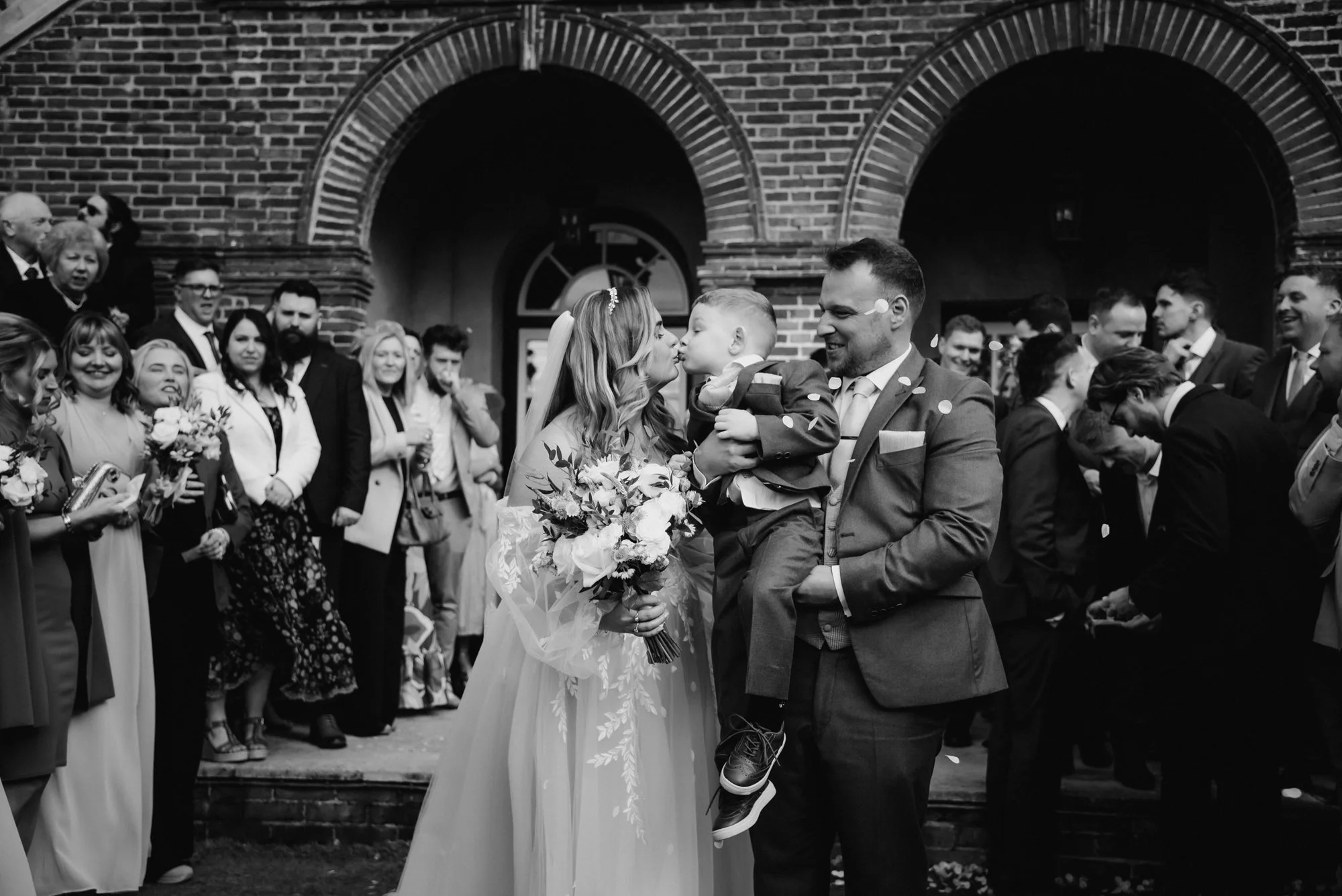 A wedding celebration with a bride, groom, and a young child outside a brick building. The bride holds a bouquet and leans in to kiss the child, who is being held by the groom. Guests surround them, some taking photos and others smiling.