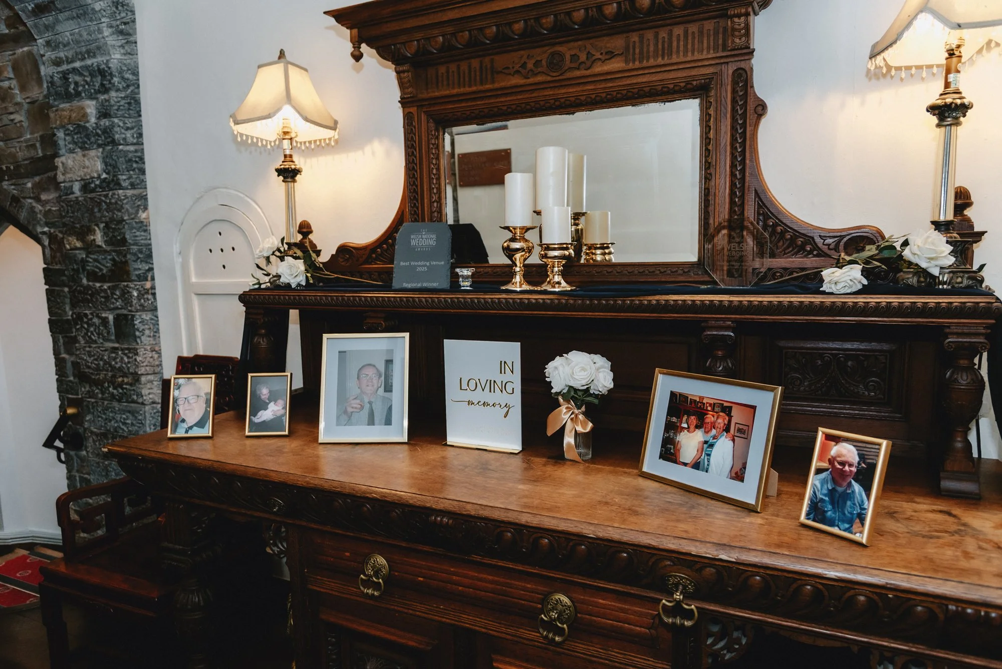 A wooden sideboard with framed photographs of elderly people, a white floral arrangement, a white card that reads 'In Loving Memory,' and candles, with a large ornate mirror and table lamps in the background.