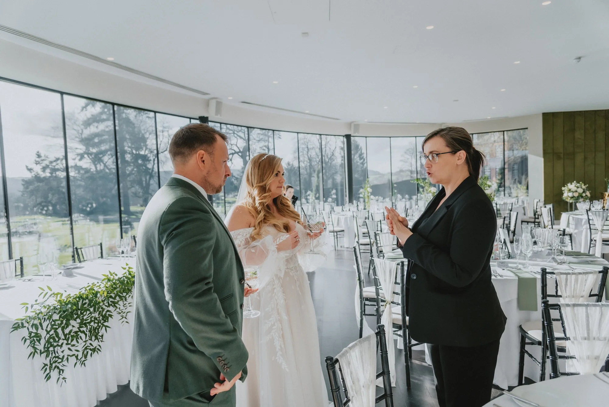 A wedding rehearsal with a bride and groom talking to an officiant in a bright, modern interior with large windows, tables set for a reception, and a scenic outdoor view.