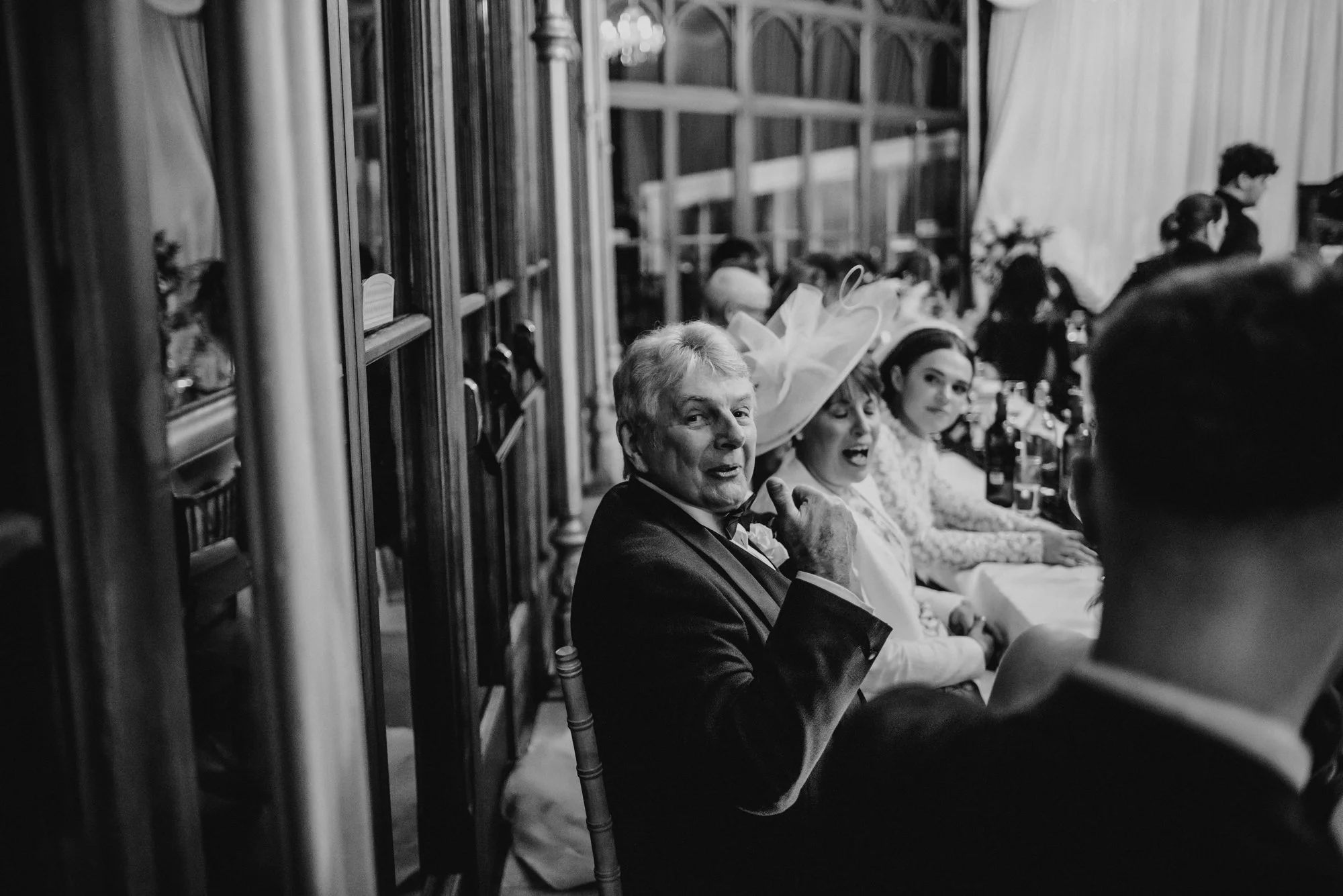 Guests sitting at a table during a formal event, with a man in a suit turning towards the camera, women next to him wearing fancy attire including a large hat, in a decorated banquet hall.