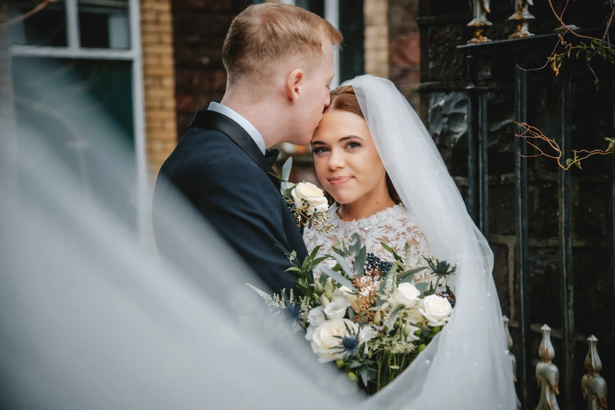 A bride and groom on their wedding day, with the groom kissing the bride's forehead and the bride holding a bouquet of white and green flowers, standing outdoors near a brick building and black iron fence.
