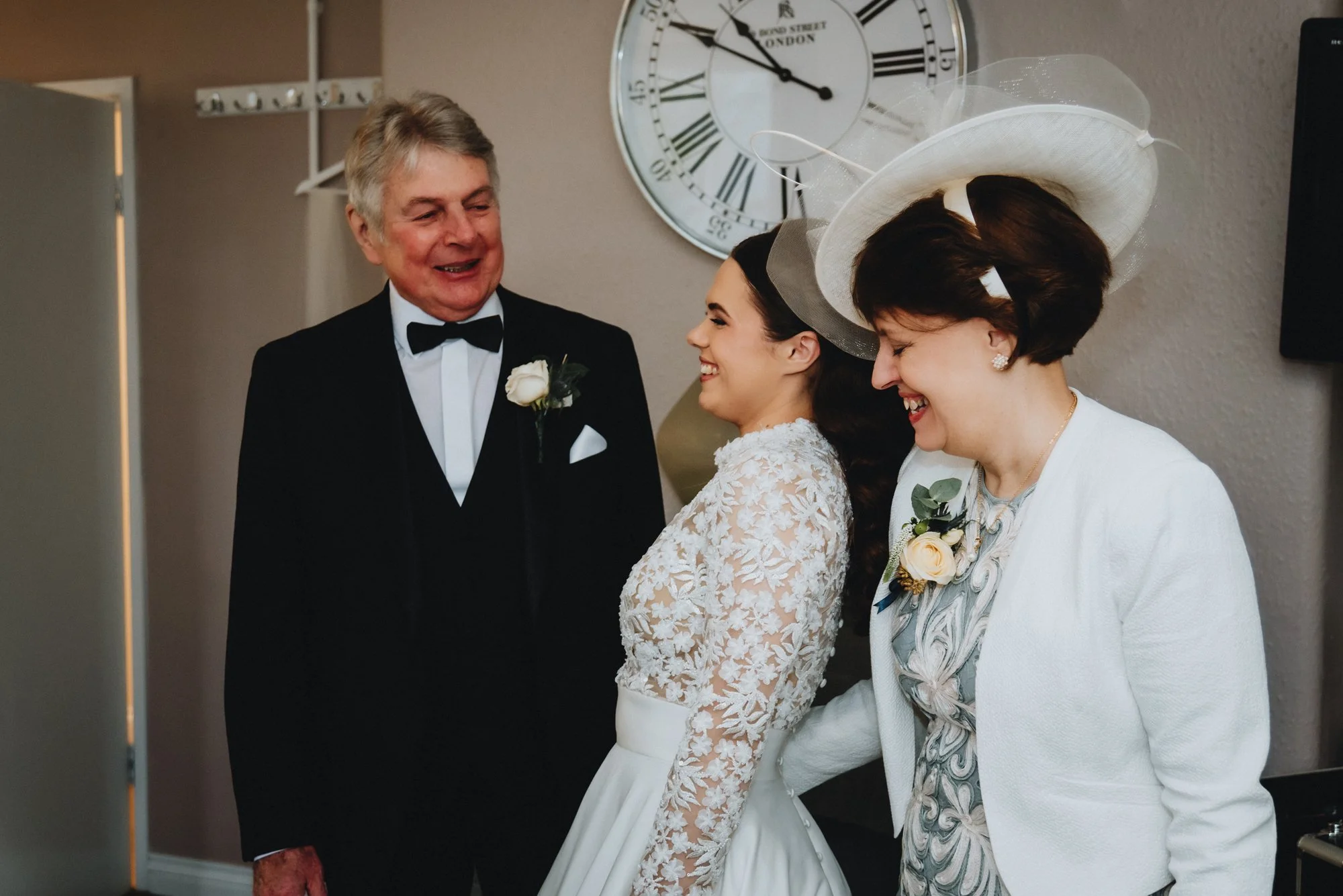 A bride in a white lace dress smiling and looking at a man in a tuxedo, accompanied by an older woman in a white outfit and large hat, both smiling.