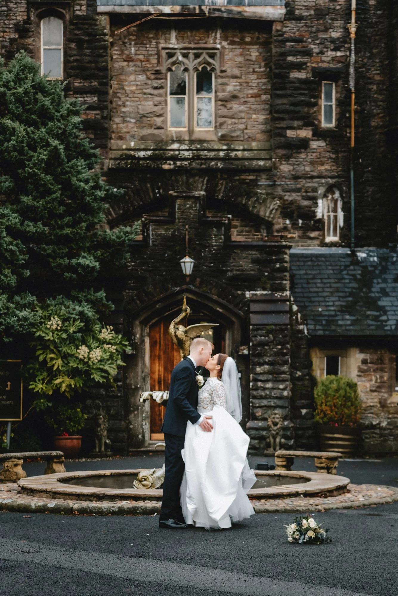 A bride and groom sharing a kiss in front of a rustic stone church with gothic windows, a wooden door, and a fountain with sculptures, including a rooster, nearby.