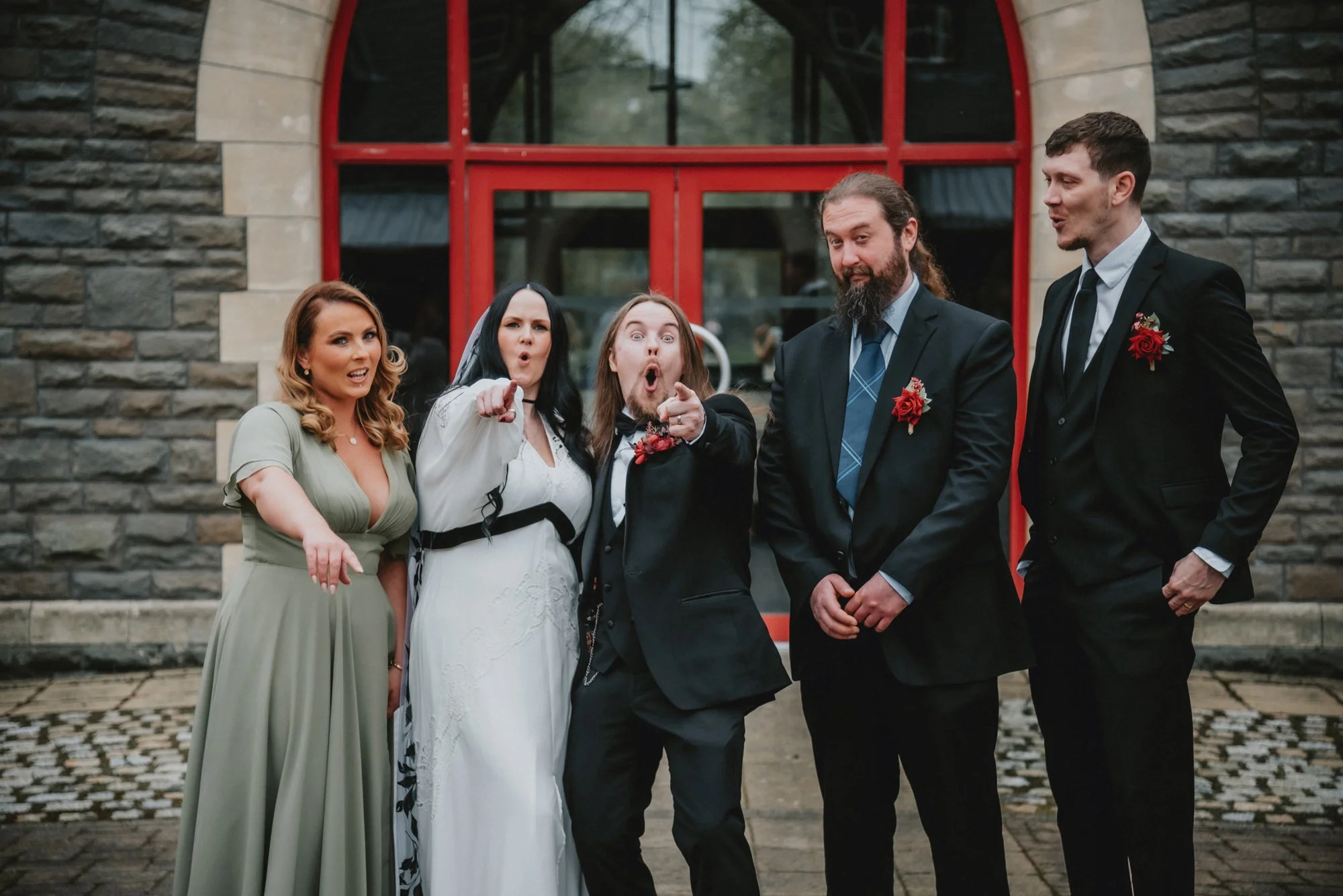 Group of five people dressed in formal attire standing outside a stone building with a red arched window behind them. They are making surprised or playful gestures, with some pointing or with their mouths open.