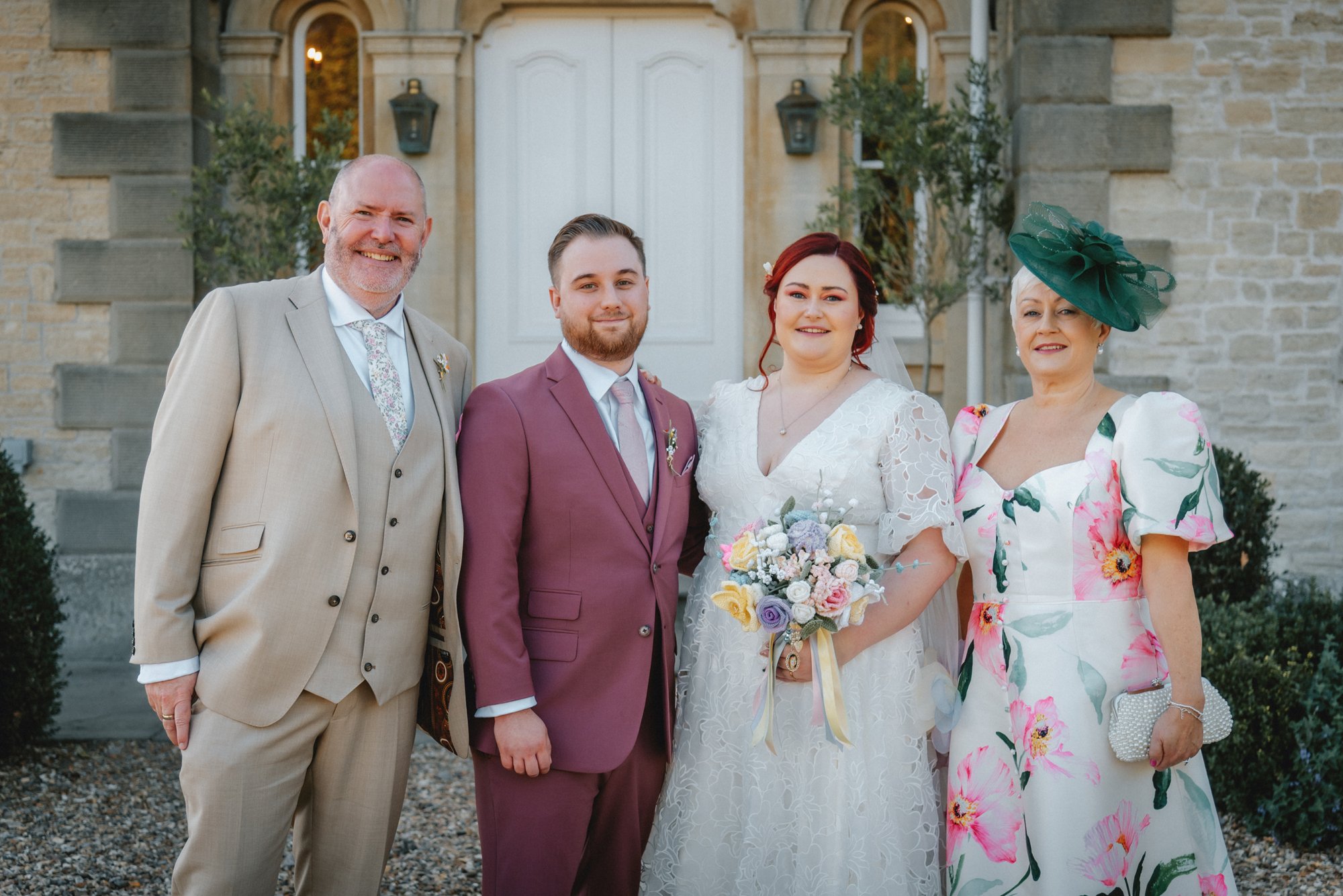 A group of five people dressed in wedding attire standing outside a stone building with a white door, smiling for a photo. The bride is holding a bouquet of pastel-colored flowers. The groom and other family members are dressed in formal suits and dr