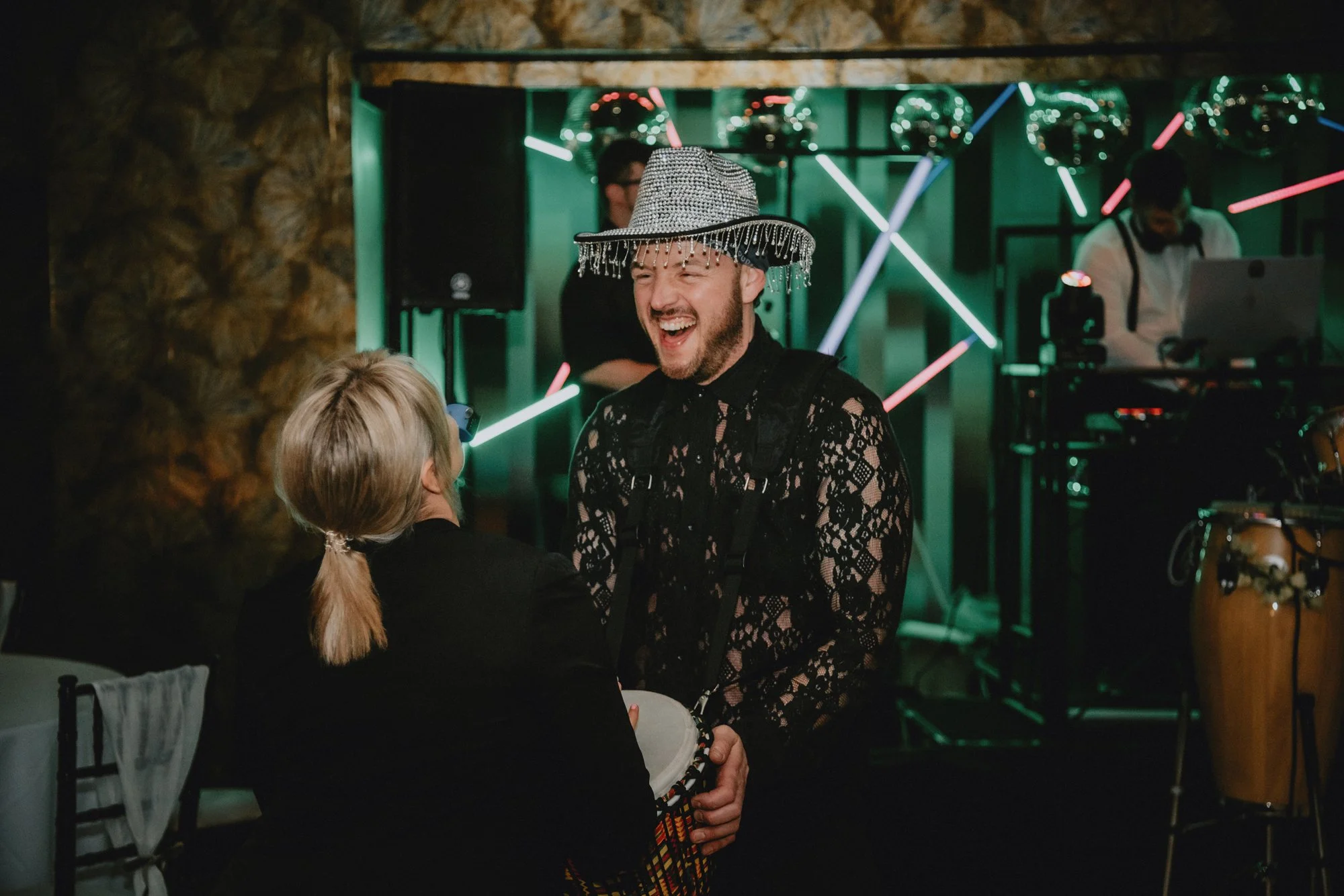 A man wearing a decorated hat and a lace shirt is laughing and holding hands with a woman with ponytail, in a festive setting with a DJ and musical instruments in the background.