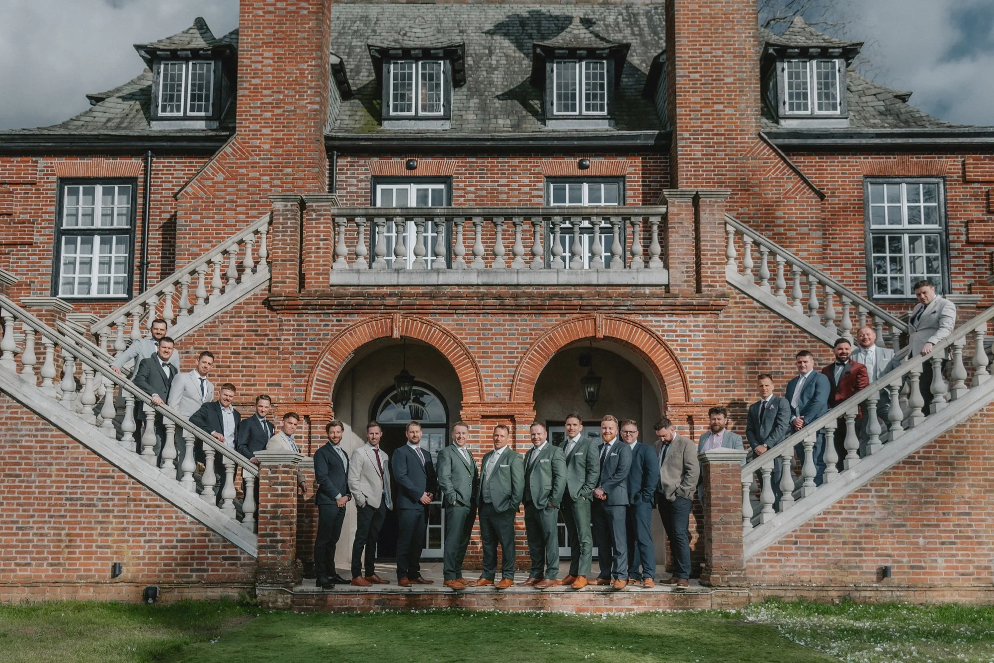 A group of men in formal suits standing on steps in front of a large brick mansion with stairs on each side, a balcony, and arched entryways.