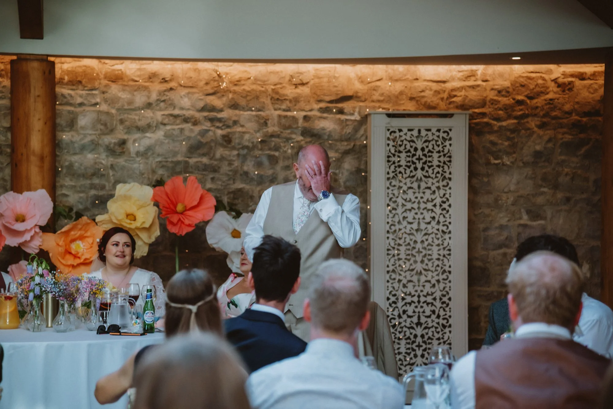 A man in a beige vest and floral tie is holding his face with one hand, standing in front of a stone wall with large paper flowers, during a wedding reception.