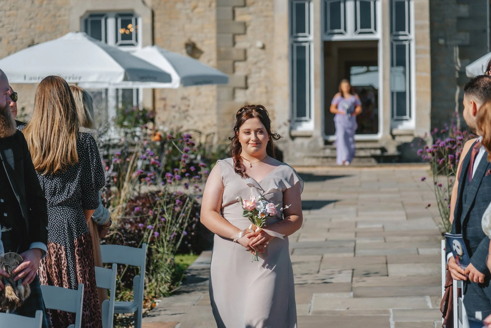 A woman in a beige dress holding a small bouquet of flowers, standing outdoors during a celebration with guests around.