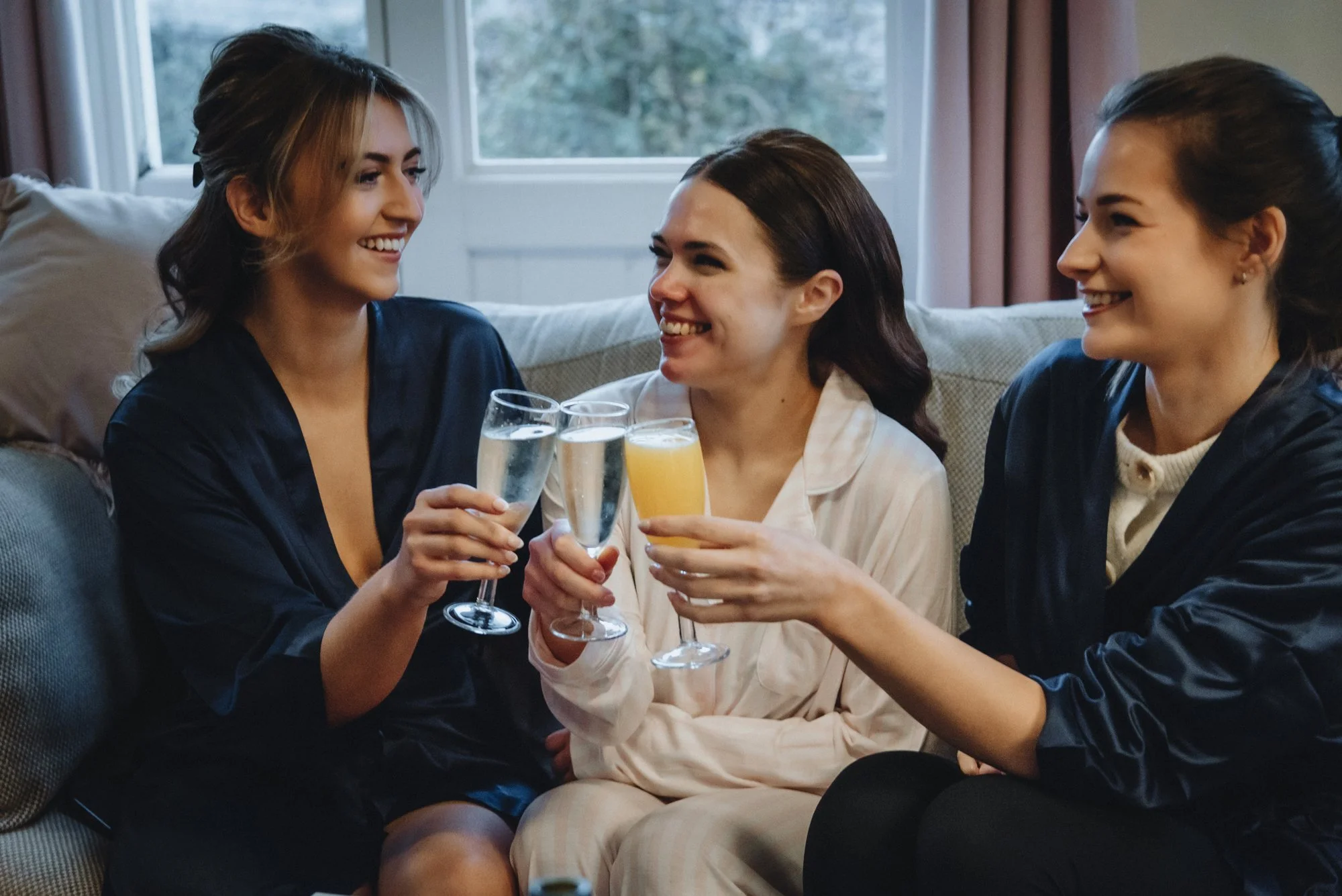 Three women sitting on a couch, smiling, and clinking glasses of champagne and orange juice during a celebration.
