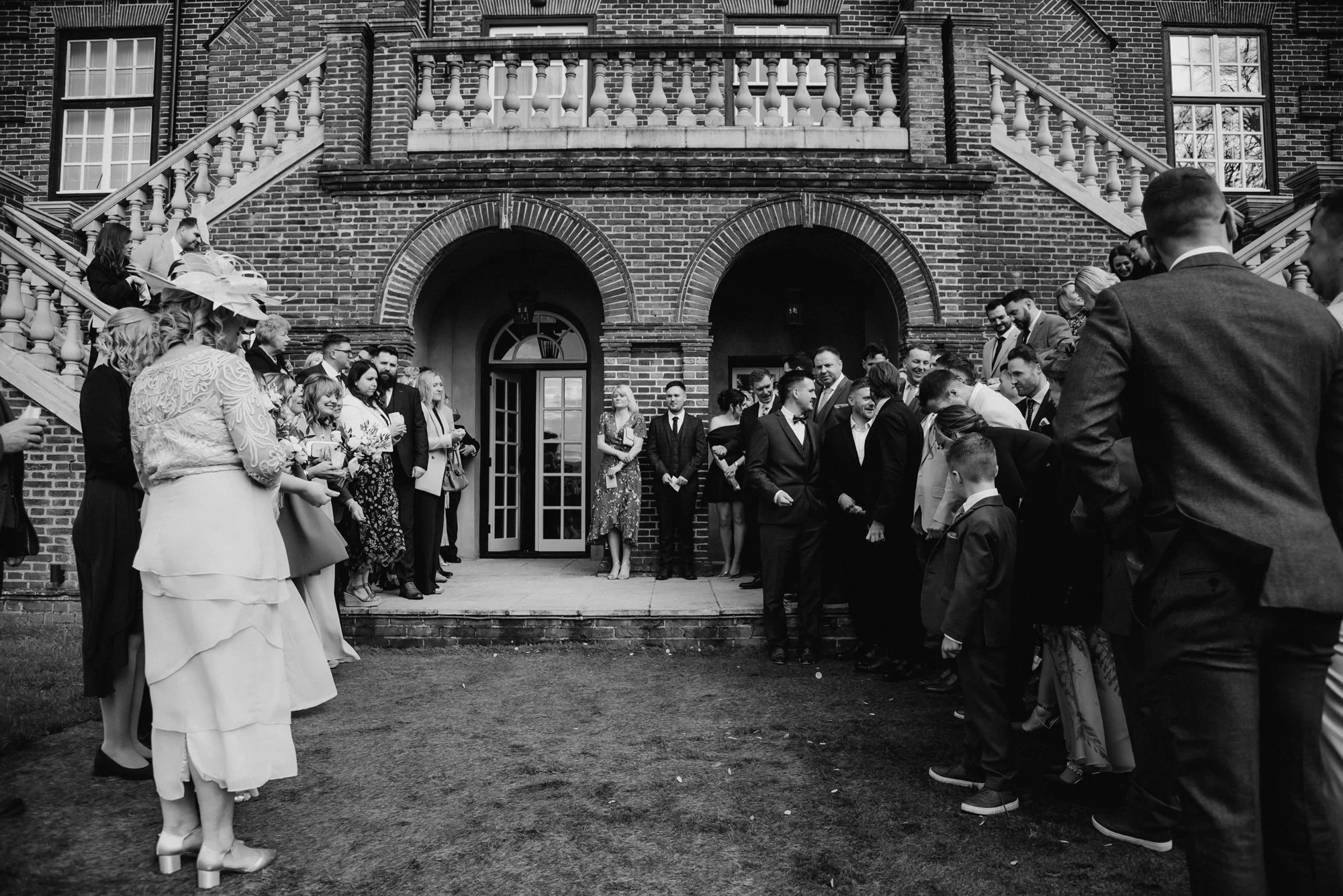 Black and white photo of a wedding gathering outside a brick building with a staircase. People are dressed formally, standing in two lines facing each other.