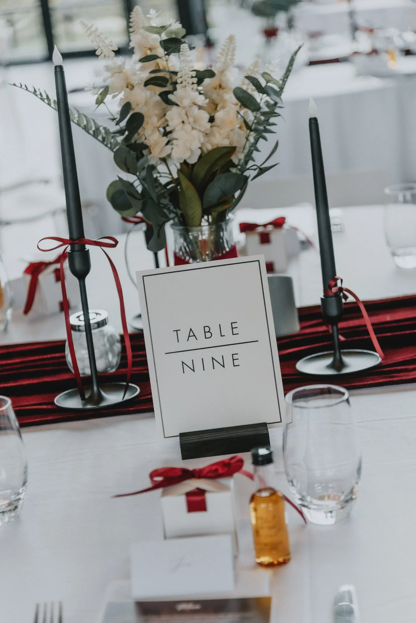 Wedding table setup with a centerpiece of white flowers, two black taper candles, and a table number sign reading 'Table Nine'. The table is decorated with a red and black striped table runner, and small gift boxes with red ribbons. Glassware, a wate