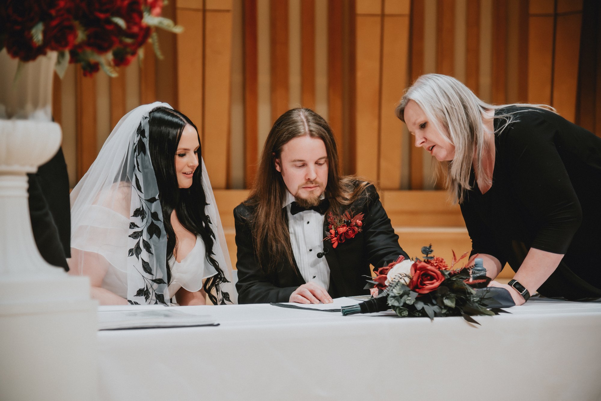 A wedding ceremony with a bride, groom, and an officiant signing a document at a table decorated with a red flower bouquet.