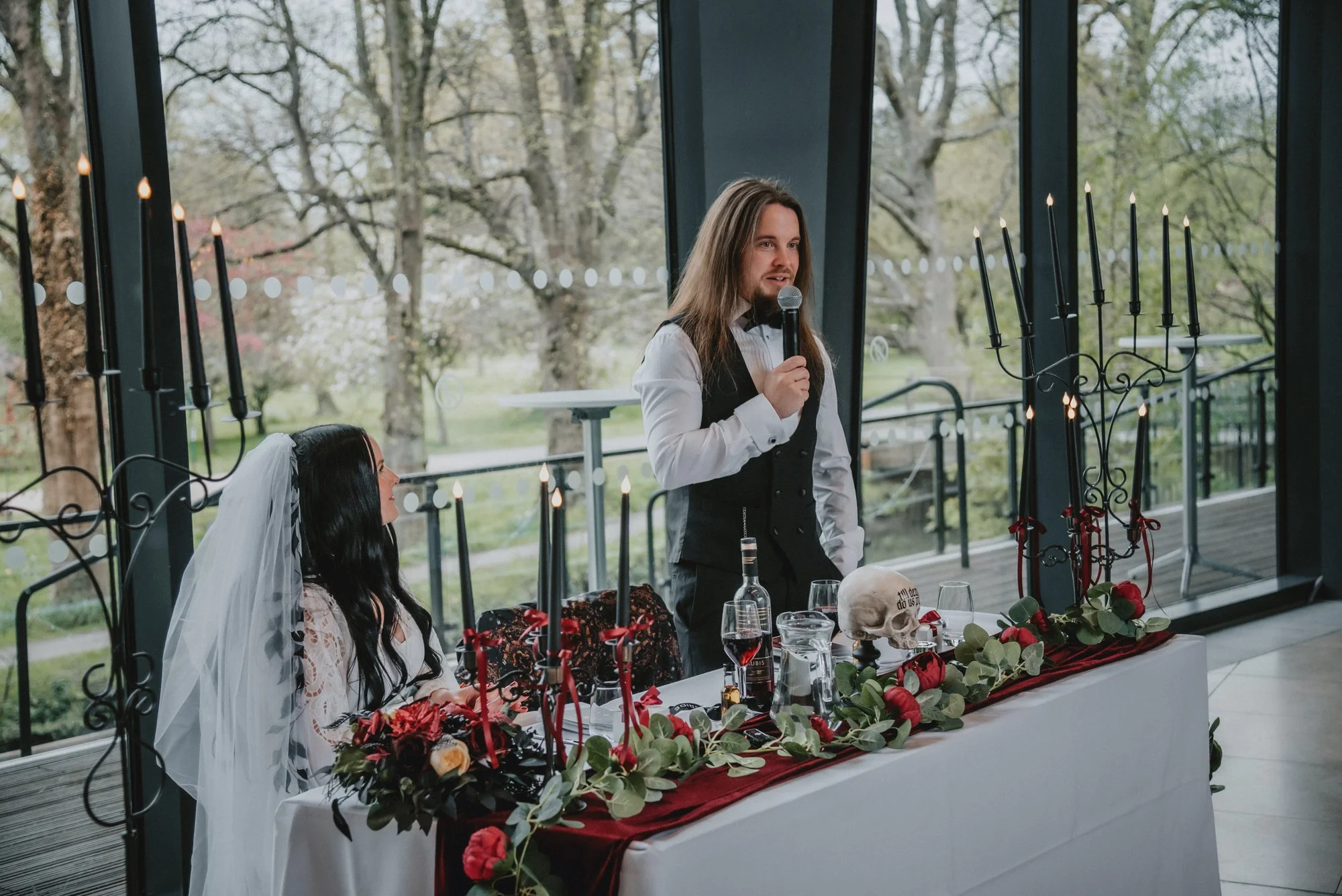 A man with long hair and a beard in a black vest and bow tie speaking into a microphone at a wedding reception. A woman with long dark hair, a white dress, and a veil sits nearby. The table has a skull, glasses of red wine, and floral decorations, an