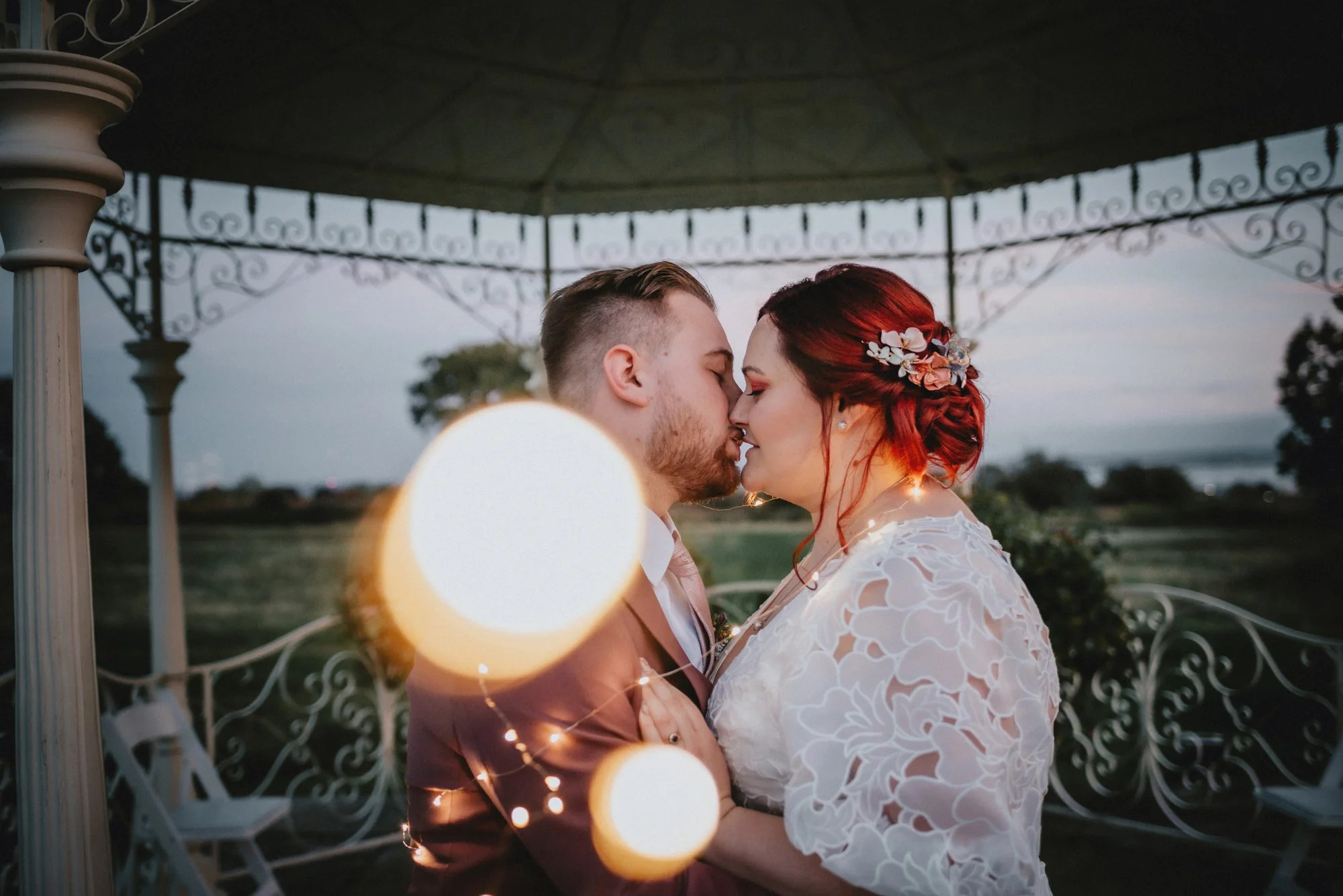 A couple kissing under a gazebo at sunset, with string lights wrapped around them, in an outdoor setting with open fields and trees in the background.