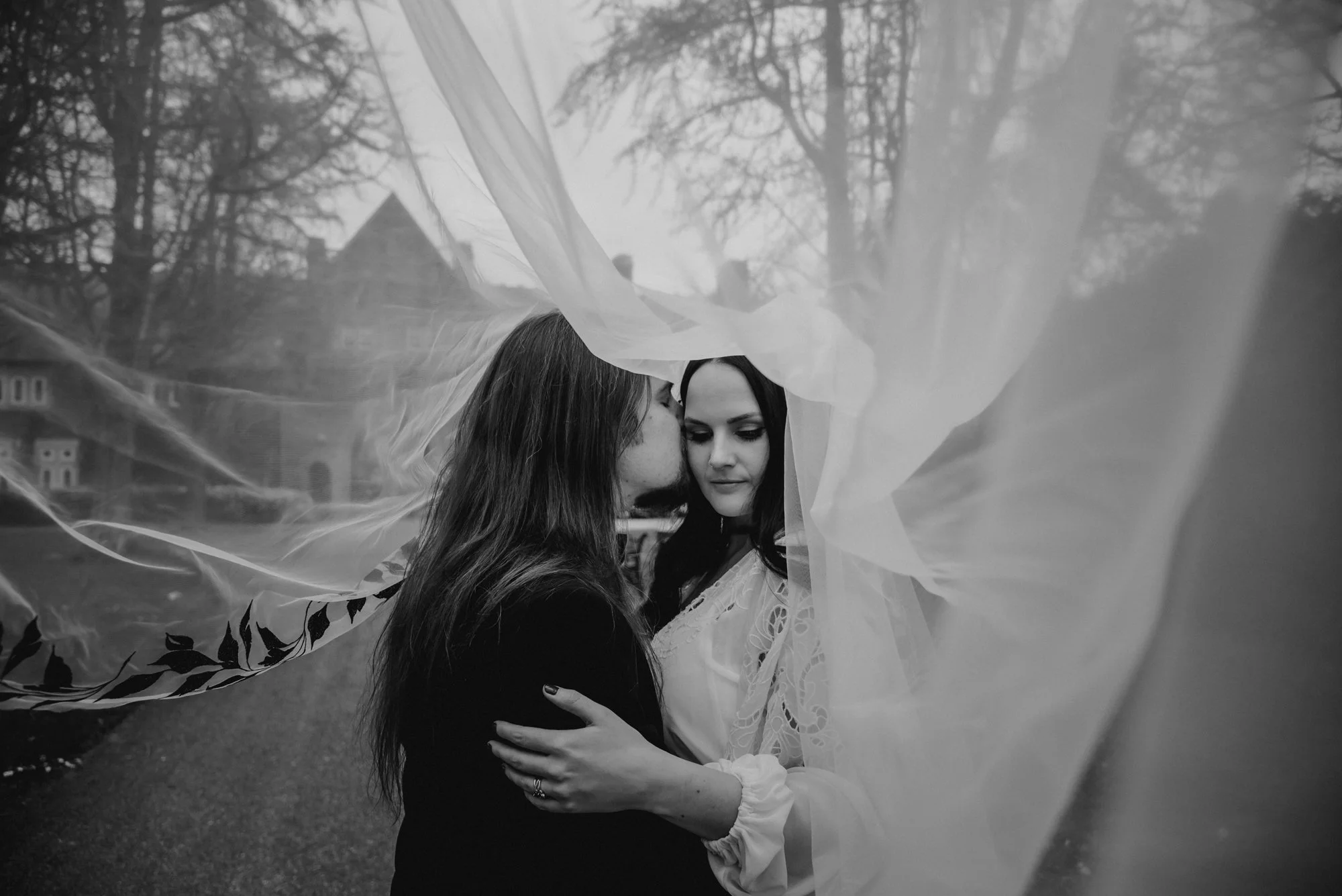 A black and white photograph of two women embracing outdoors, with one woman whispering into the other's ear. A sheer fabric or veil surrounds them, creating a dreamy atmosphere. In the background, there are trees and a house.