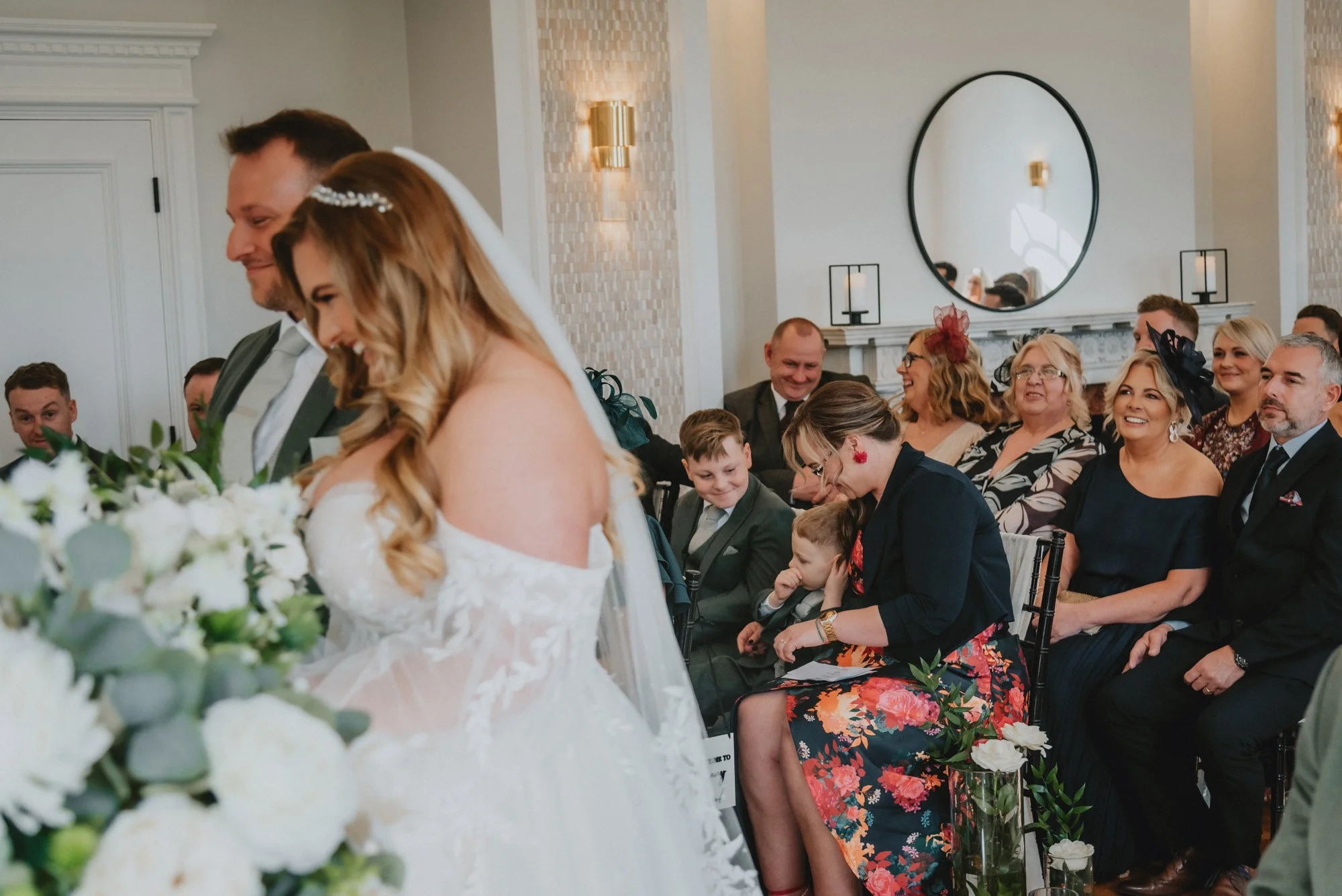 Wedding ceremony with the bride and groom standing together, guests seated and smiling, some children among the guests, in a bright and elegant room.