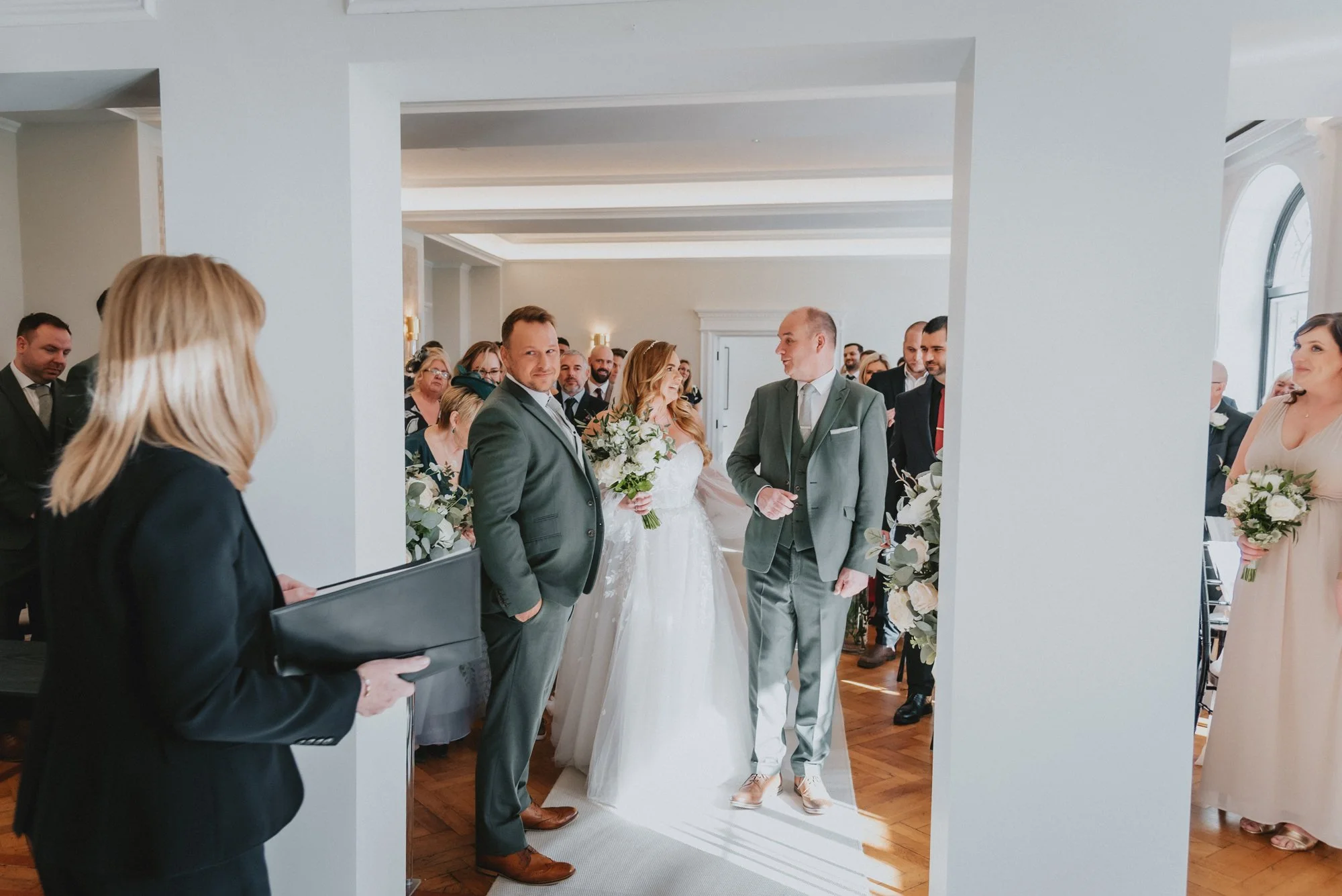 A wedding ceremony with the bride in a white dress holding a bouquet of flowers, flanked by two men in suits, one on each side, inside a bright, decorated room with multiple guests.